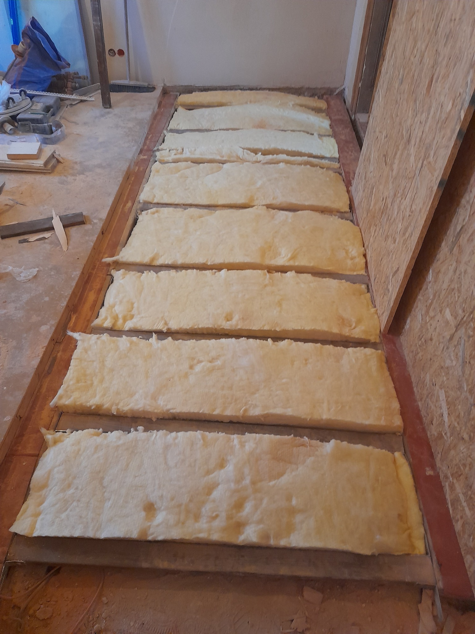 Izolacja podłogi z wełny mineralnej w trakcie prac wykończeniowych. Widoczne fragmenty konstrukcji podłogi i ściany z płyty OSB. Ujęcie z góry, w trakcie realizacji.