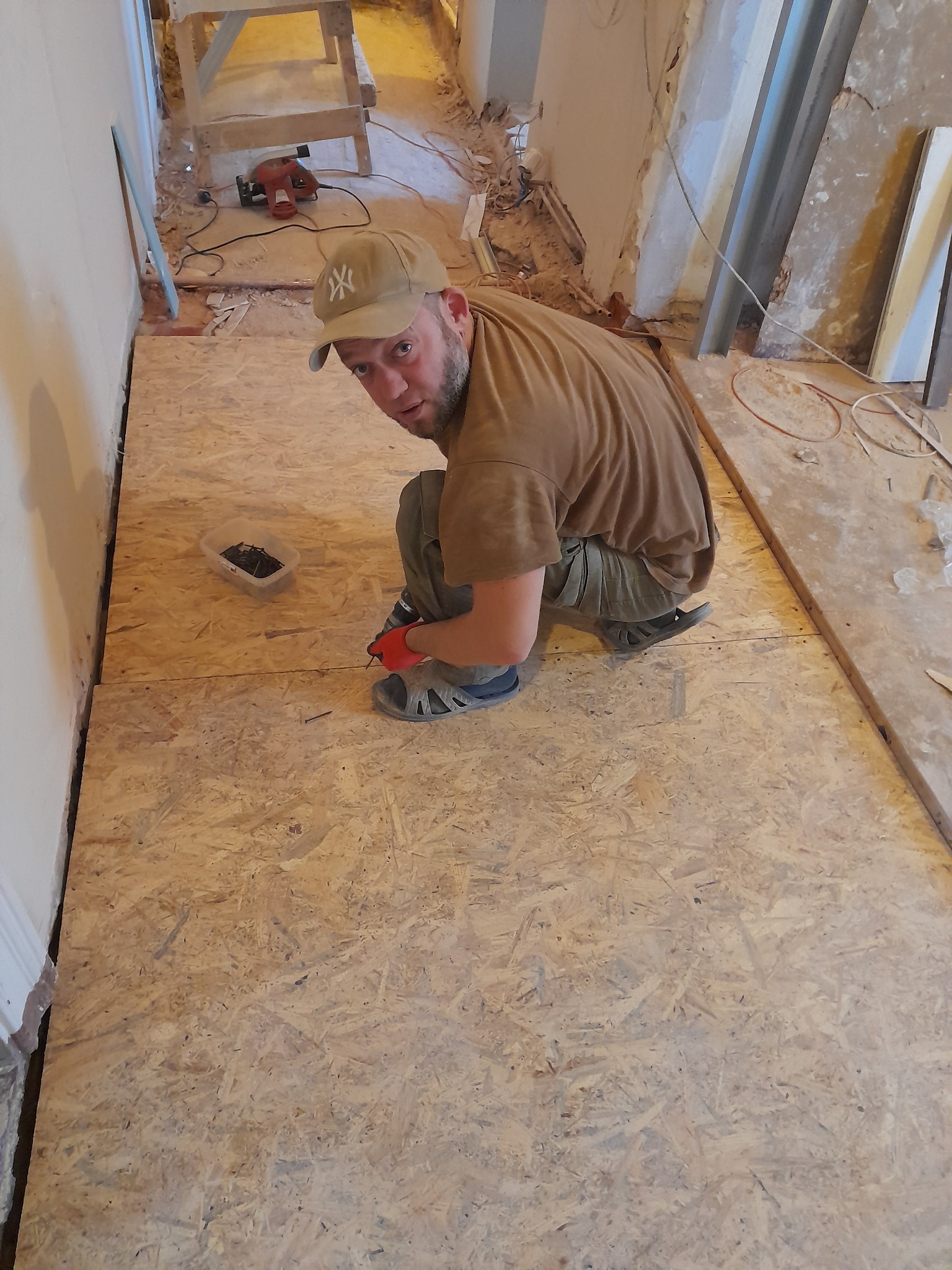 Montaż podłogi z płyty OSB w korytarzu podczas remontu. Widoczny pracownik w trakcie pracy z wkrętarką i pudełkiem wkrętów. W tle narzędzia i elementy konstrukcyjne.