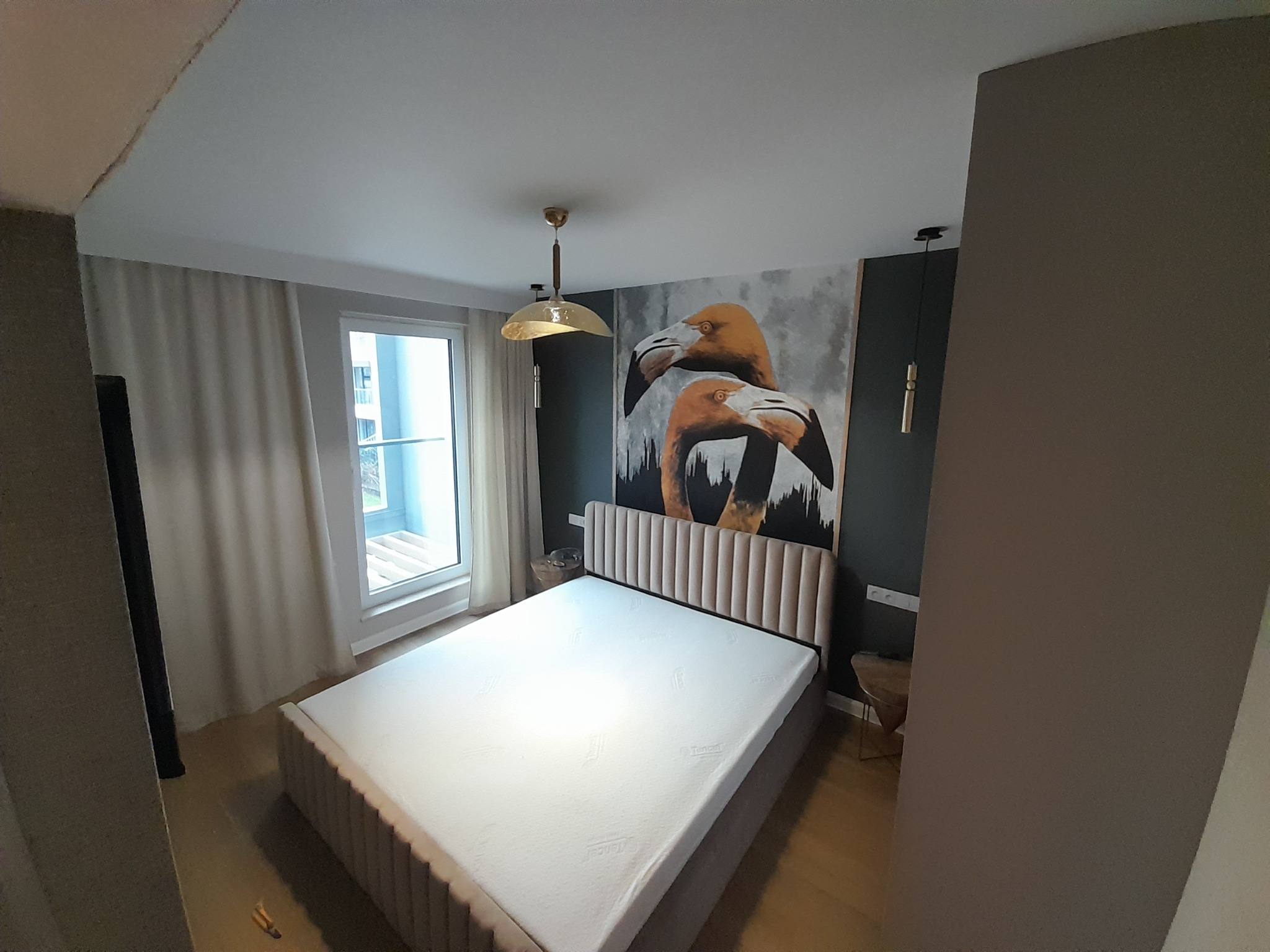 Nowoczesna sypialnia z łóżkiem w beżowej tapicerce, obrazem flamingów na ścianie i złotą lampą. Minimalistyczny design, jasne zasłony i drewniana podłoga.