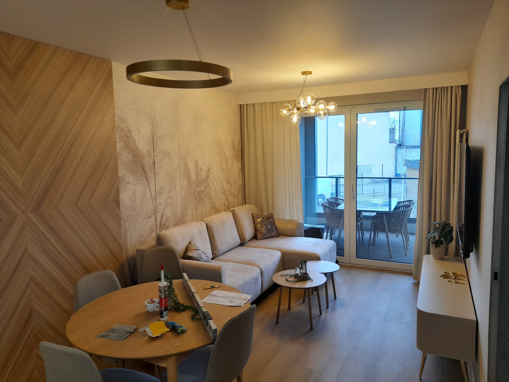 Jasne wnętrze salonu z drewnianą ścianą, beżową sofą i okrągłym stołem. Widoczne narzędzia sugerują prace wykończeniowe. Dwa nowoczesne żyrandole.