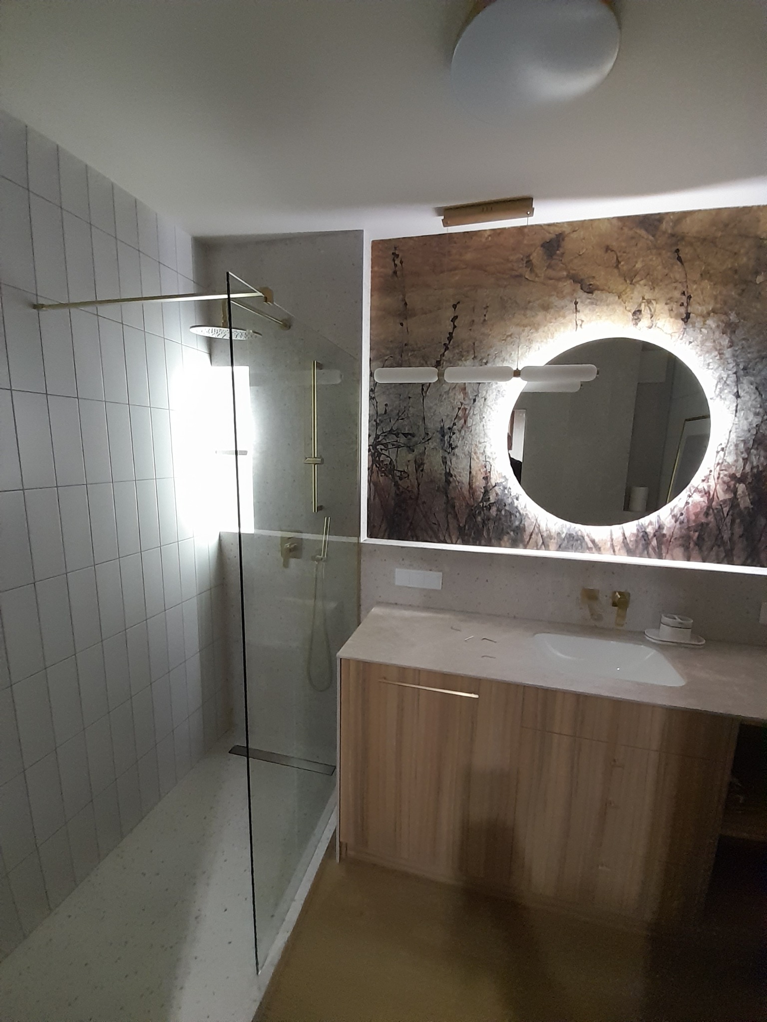 Nowoczesna łazienka z kabiną prysznicową typu walk-in, umywalką w szafce z drewnianym fornirem i okrągłym lustrem z podświetleniem LED na dekoracyjnej ścianie.