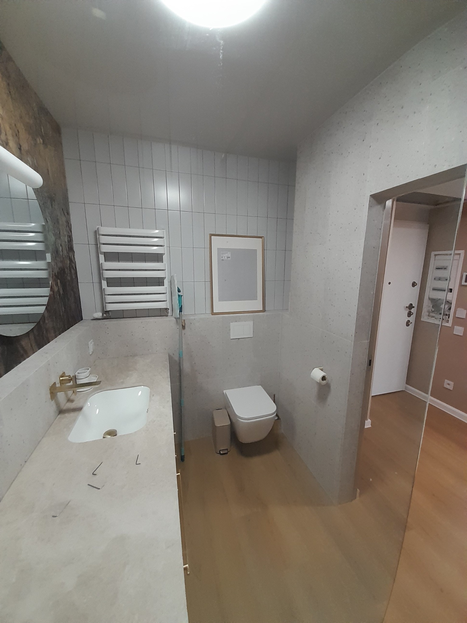 Jasna łazienka po remoncie: biała umywalka z złotą armaturą, toaleta, grzejnik drabinkowy i lustro z drewnianym akcentem. Minimalistyczny design z drewnianą podłogą.