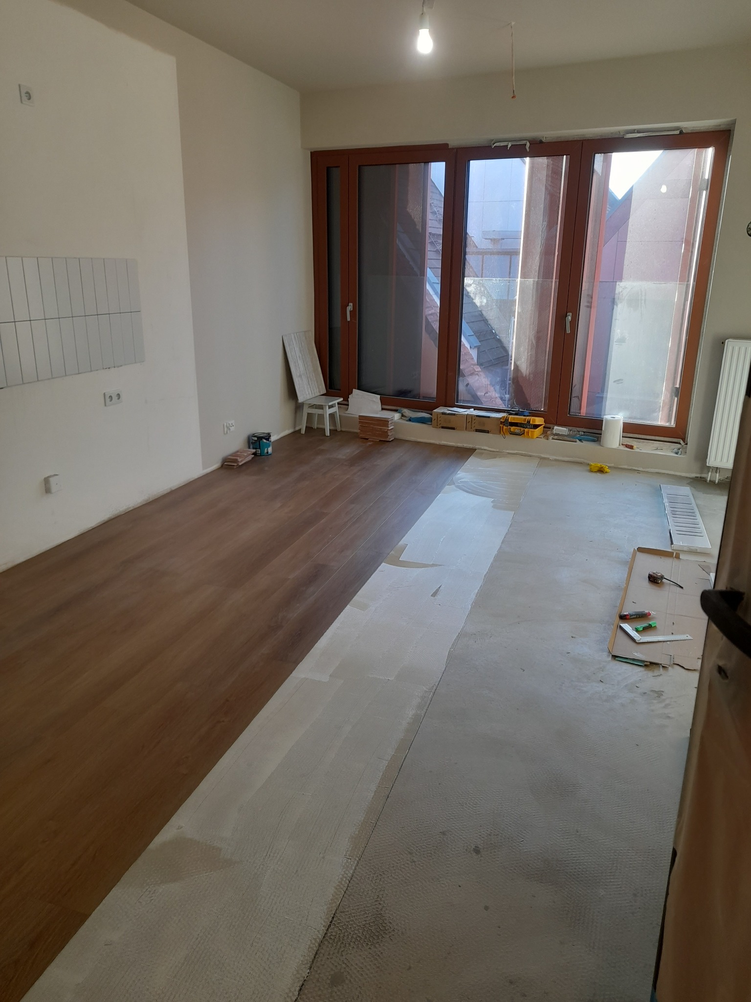 Układanie paneli podłogowych w jasnym pokoju z dużym oknem. Część podłogi wyłożona, druga przygotowana pod wylewkę. Widoczne narzędzia i materiały budowlane.