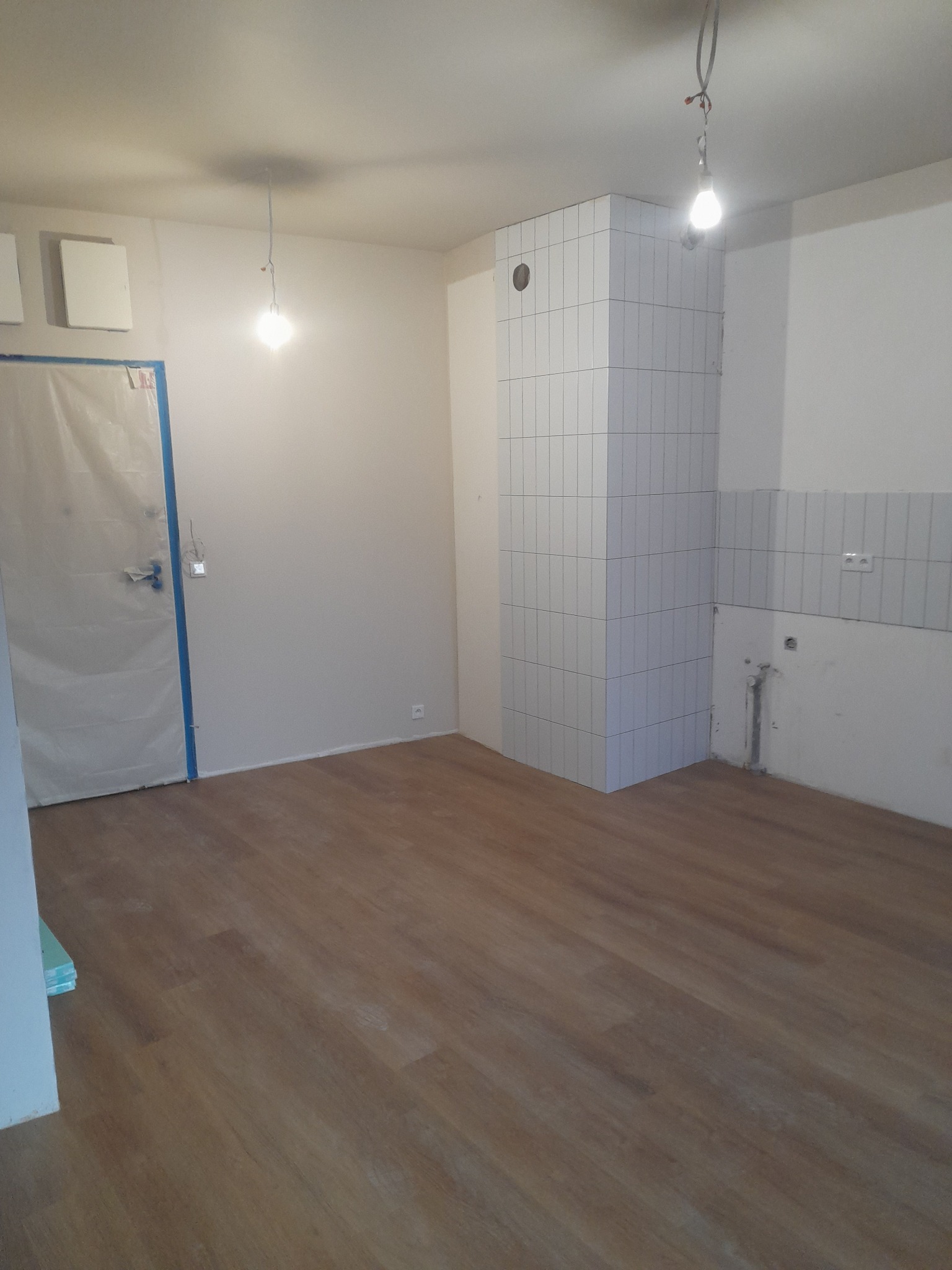 Wnętrze kuchni w trakcie prac wykończeniowych: ułożona podłoga, częściowo położone płytki na ścianie i obudowie pionu, zabezpieczone drzwi folią malarską.