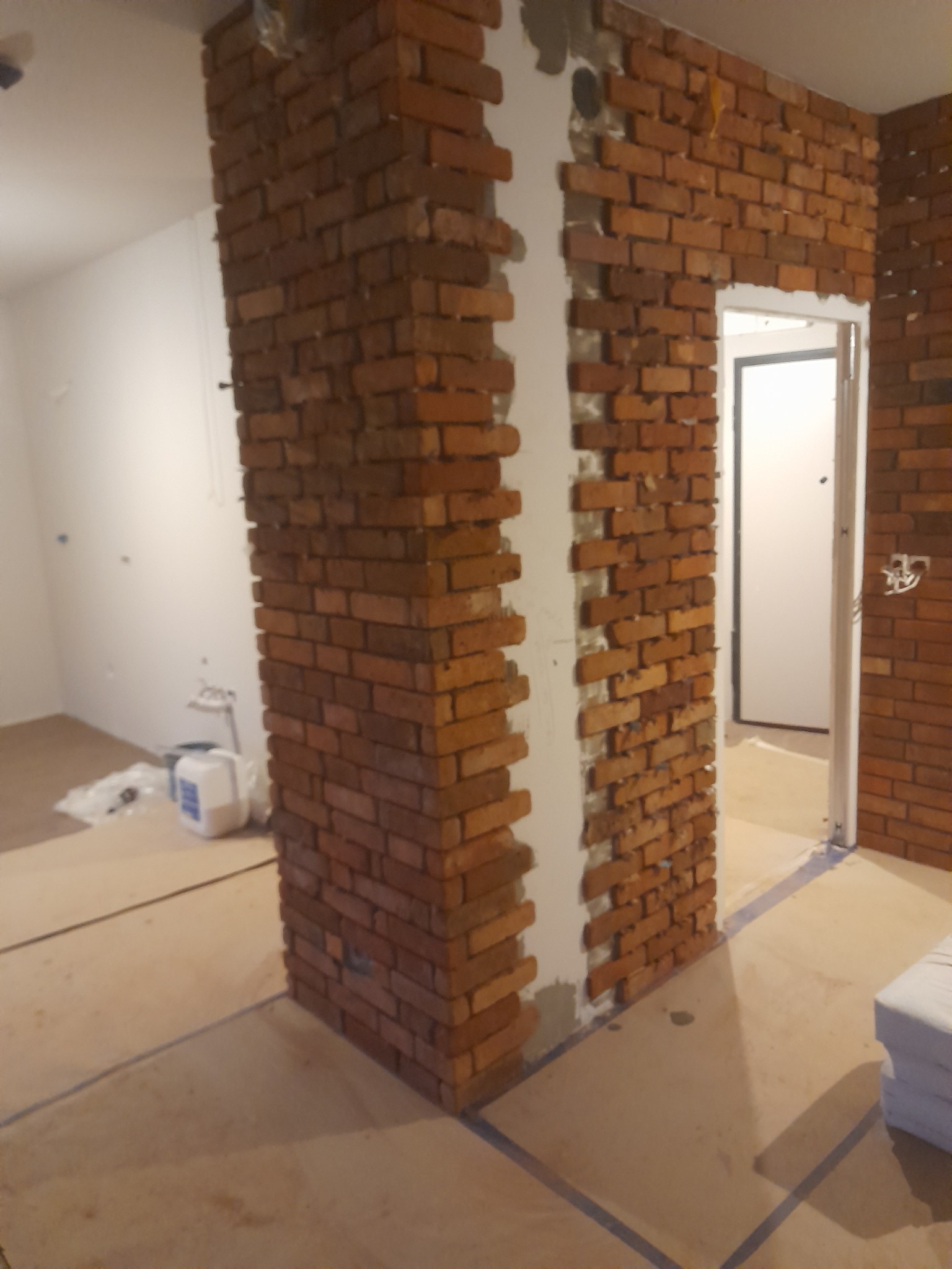 Ceglana ściana dekoracyjna w trakcie montażu, widoczne fragmenty surowej ściany i białe fugi. W tle widoczne materiały budowlane i otwarte drzwi.