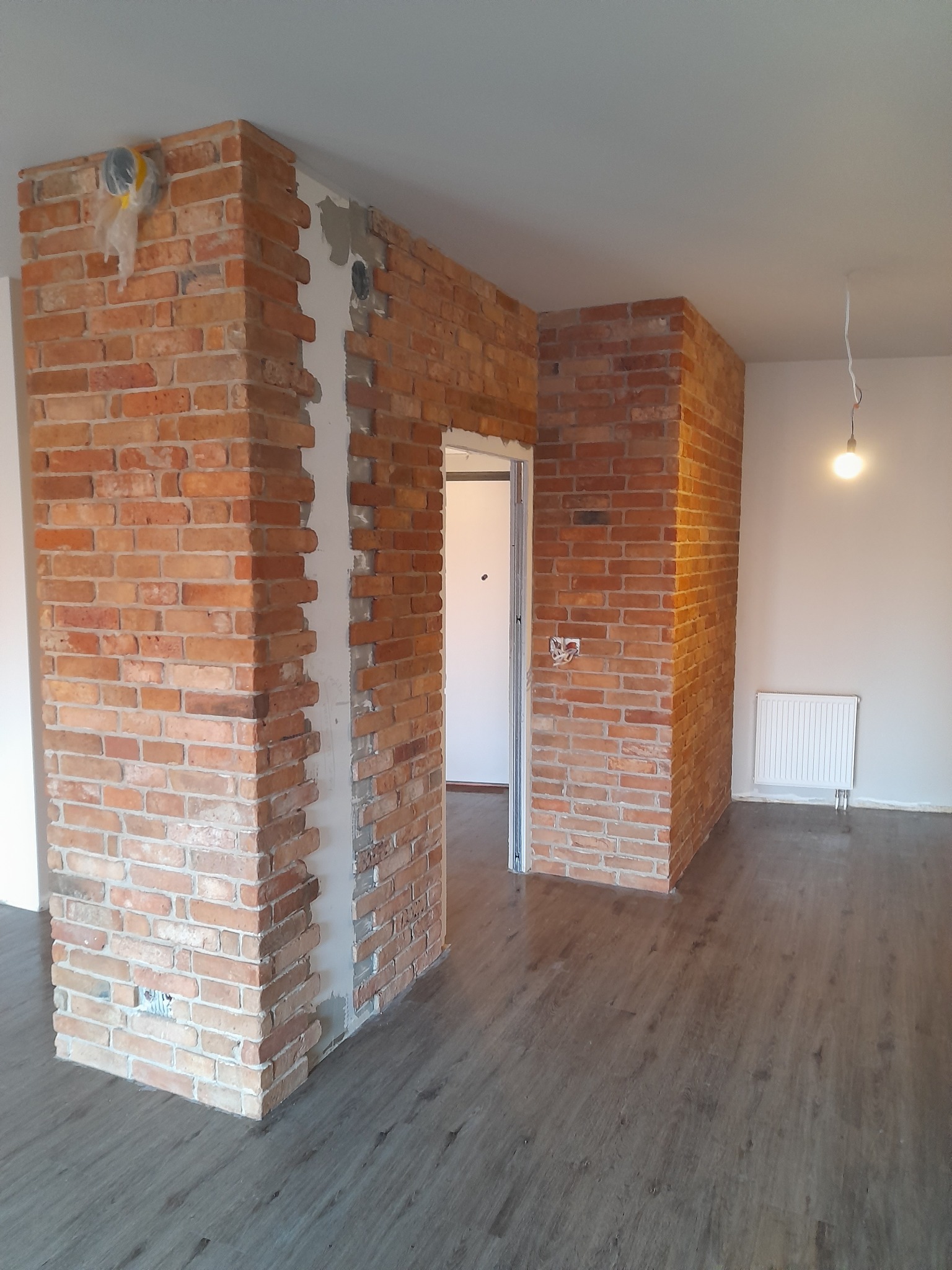 Ceglana ściana działowa z widocznymi fragmentami instalacji elektrycznej, surowy styl, drewnopodobna podłoga, grzejnik. Element wykończenia wnętrza w trakcie prac.
