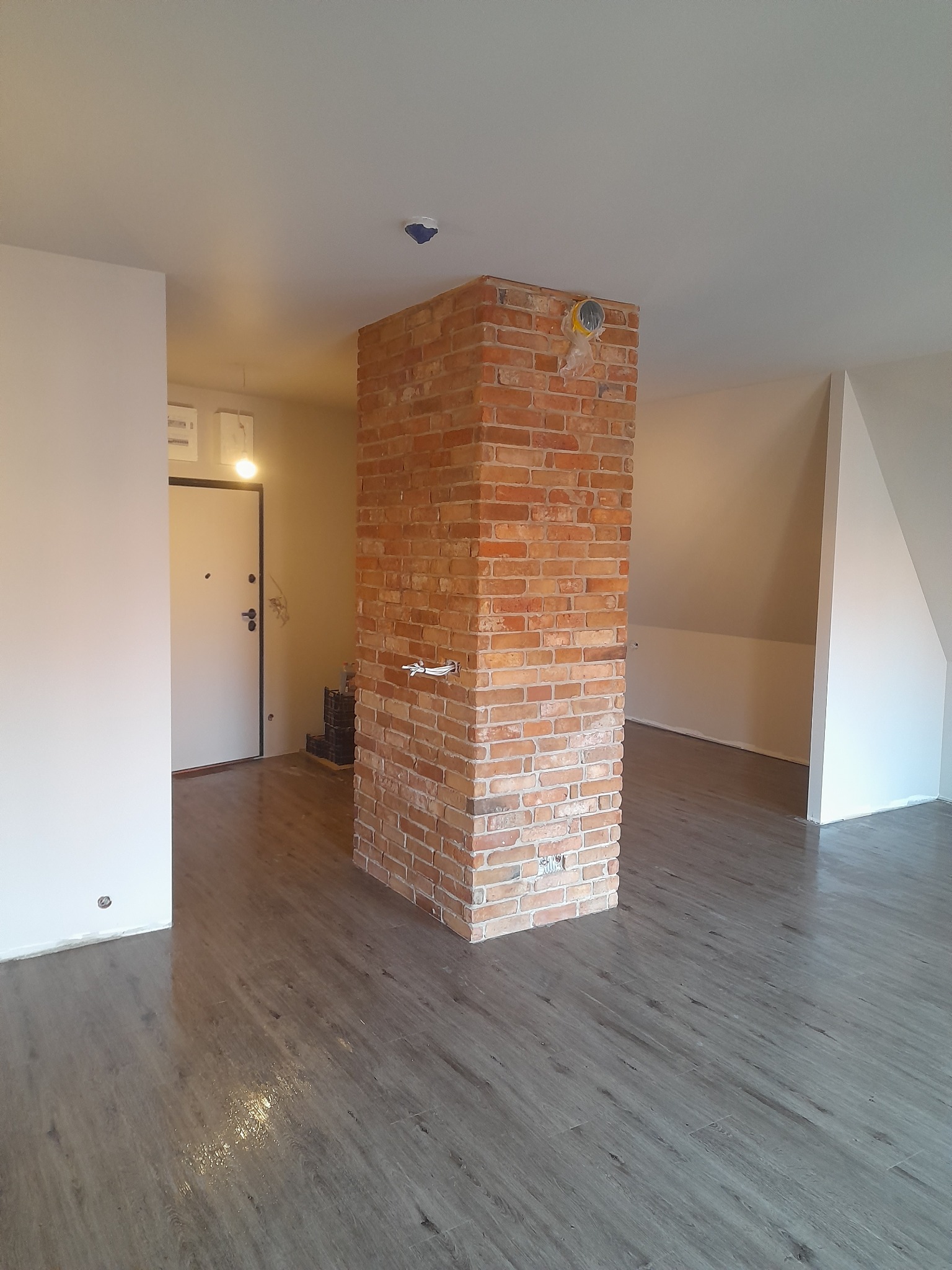 Wykończone wnętrze z kolumną z czerwonej cegły i panelami podłogowymi w odcieniach szarości. Widoczne drzwi wejściowe i fragment poddasza. Oświetlenie sufitowe.