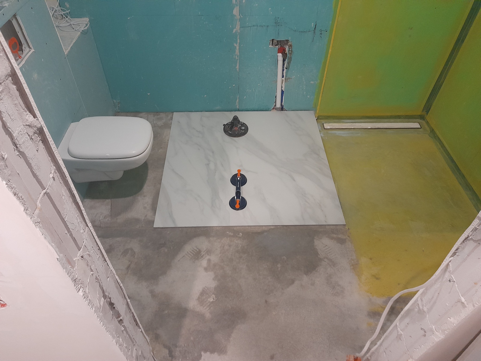 Wnętrze łazienki w trakcie prac wykończeniowych: ułożona płyta podłogowa z marmurowym wzorem, widoczne narzędzia glazurnicze, ściany przygotowane pod dalsze prace.