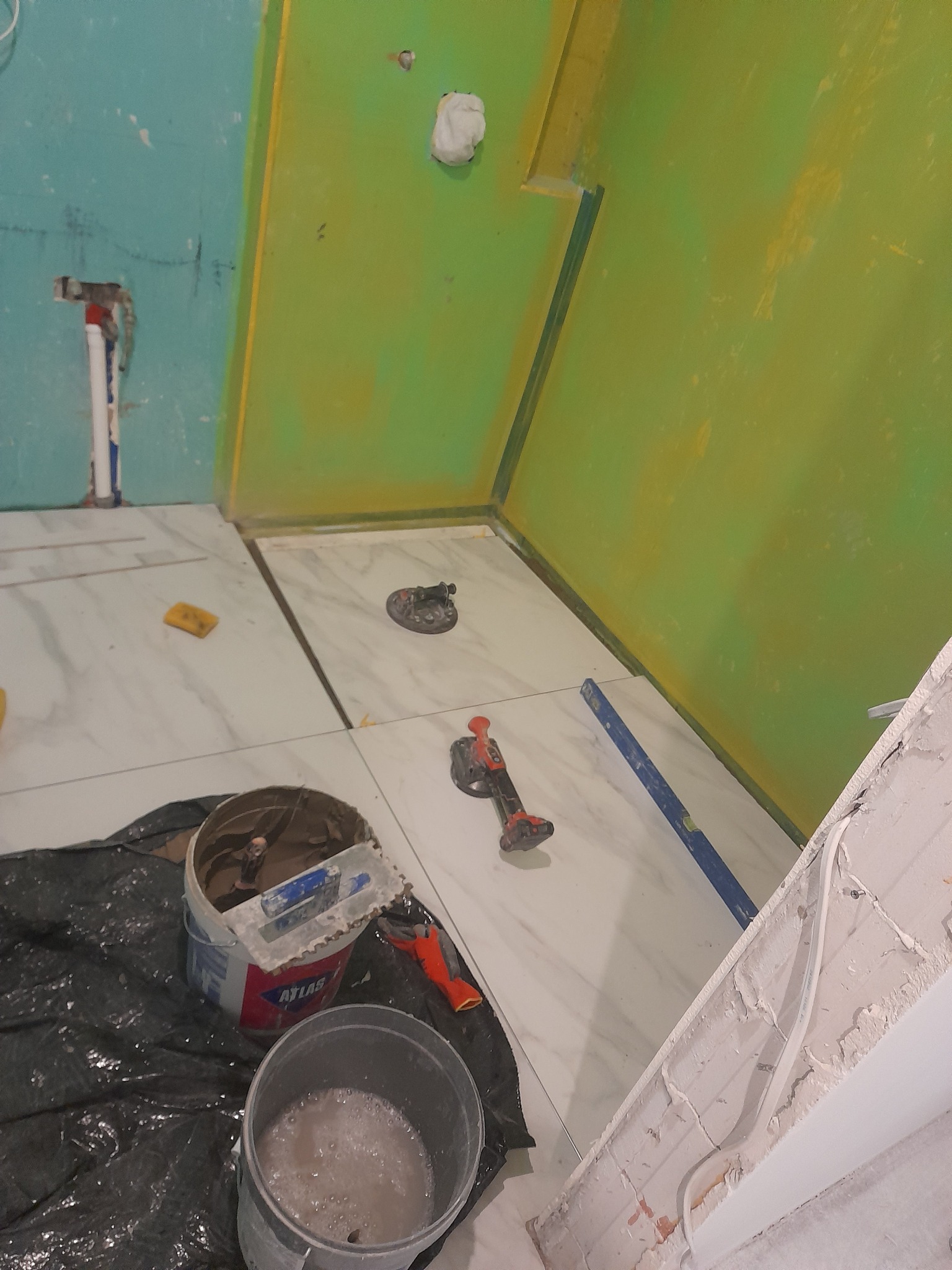 Układanie białych płytek imitujących marmur w kabinie prysznicowej. Widoczne narzędzia glazurnicze: poziomica, przyssawki, wiadro z klejem Atlas i zaprawą.