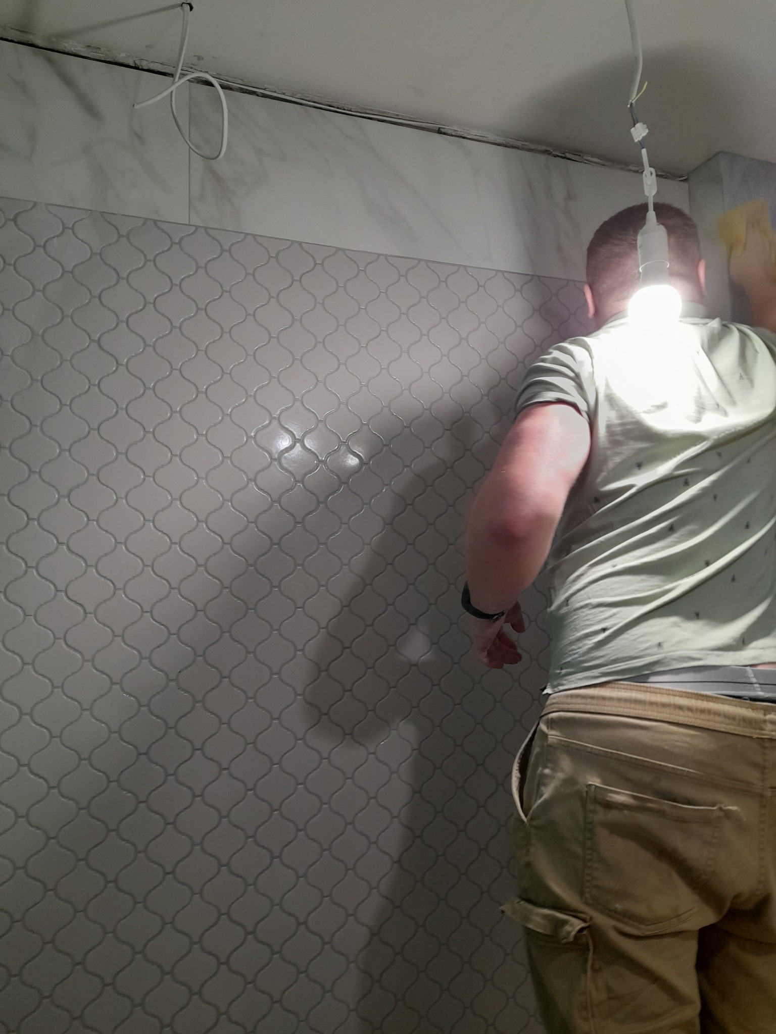 Glazurnik układa mozaikę w łazience, oświetlając sobie pracę wiszącą żarówką. Widoczny fragment ściany z ułożoną mozaiką i fragment sylwetki glazurnika.