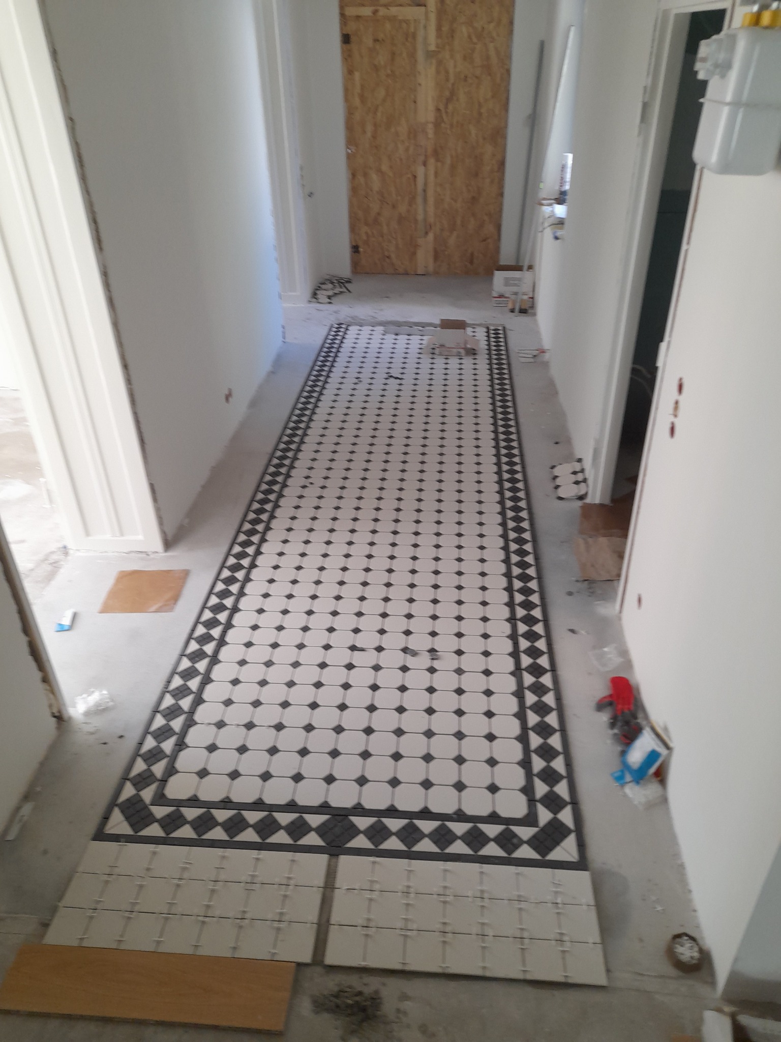 Układanie dekoracyjnej, czarno-białej mozaiki podłogowej w korytarzu. Częściowo ułożona podłoga z widocznymi płytkami do dalszego montażu. Białe ściany i drzwi w tle.