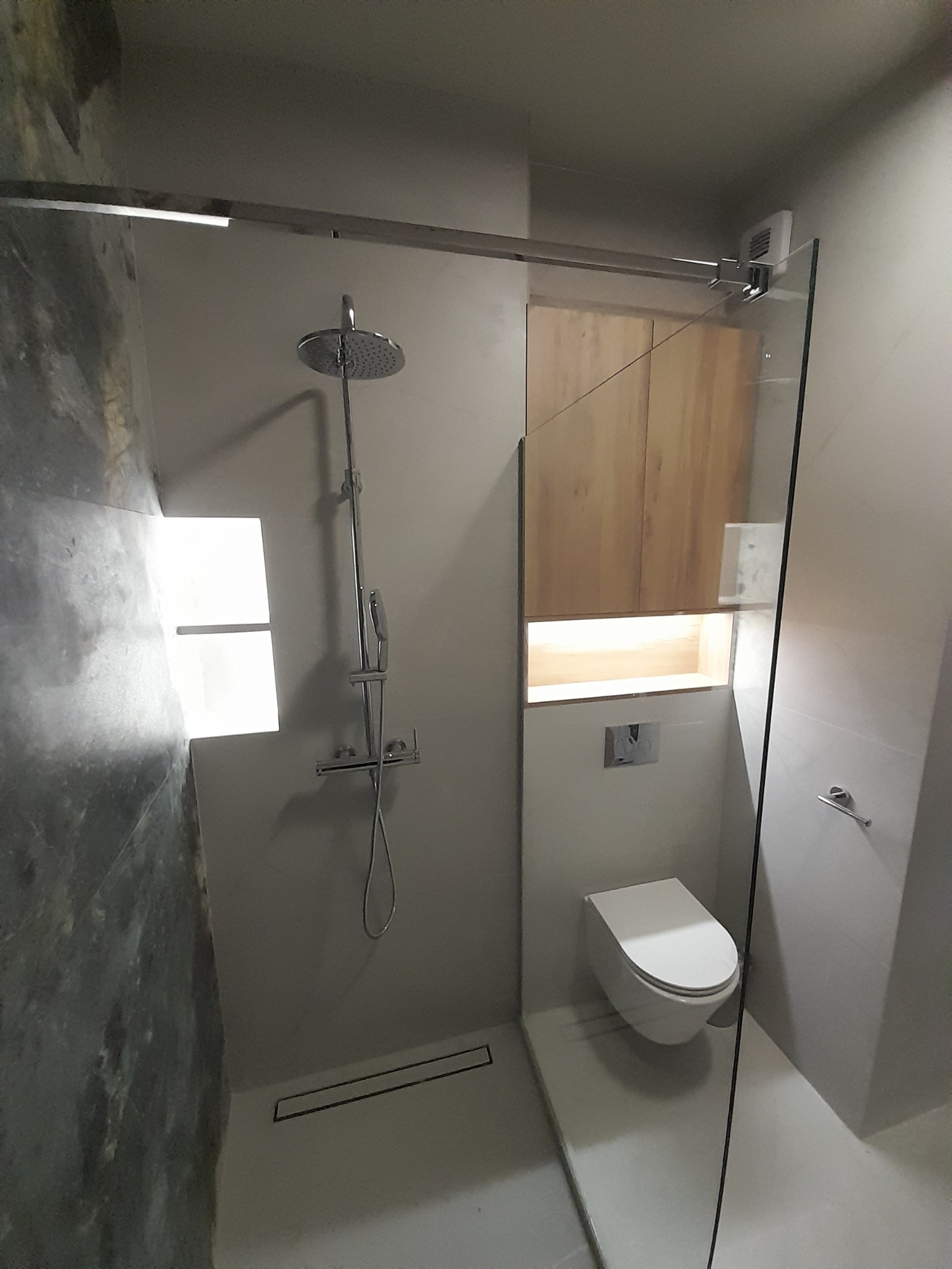 Nowoczesna łazienka z kabiną prysznicową typu walk-in, podwieszaną toaletą i drewnianą szafką z podświetleniem. Minimalistyczny design, szare płytki i szklana ściana.