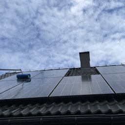 ABMH Andrzej Pawelec - Czyszczenie paneli fotowoltaicznych na dachu domu za pomocą szczotki teleskopowej. Widoczne panele, dachówka i fragment nieba z chmurami.