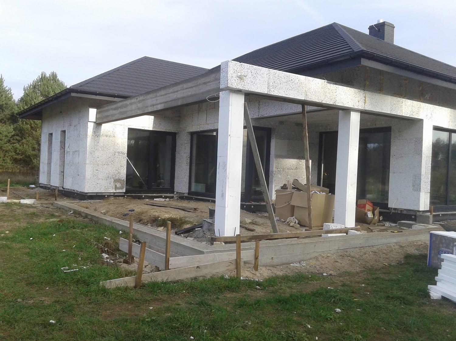 Budowa domu jednorodzinnego z białego bloczka, widoczne fundamenty i zadaszenie tarasu w trakcie realizacji, czarne okna, dach z dachówki.