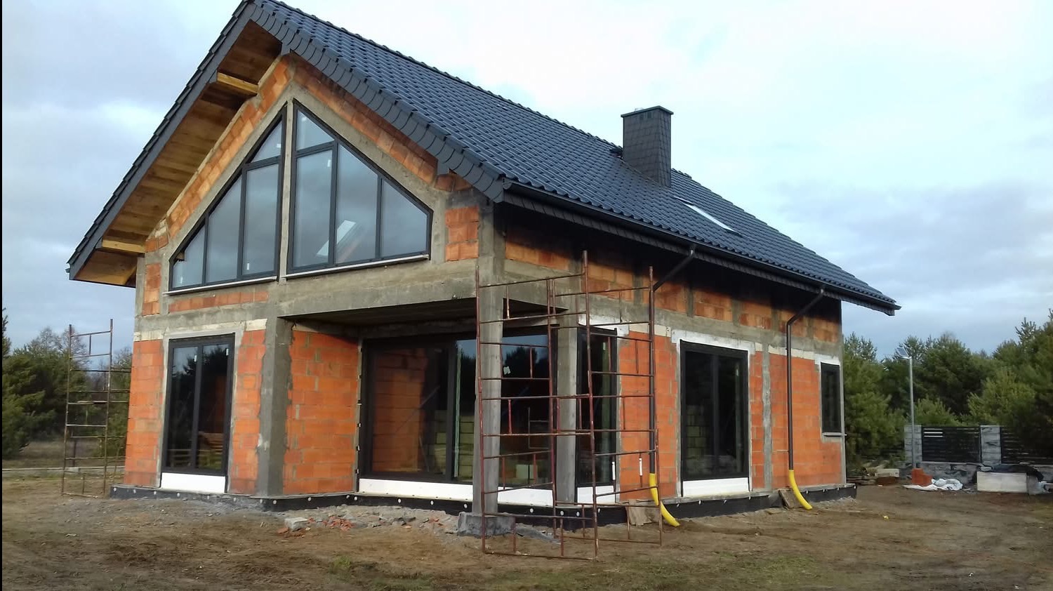 Surowy stan domu z cegły z czarną dachówką i dużymi oknami, widoczne rusztowania i przygotowania do elewacji. Charakterystyczne trójkątne okno na poddaszu.