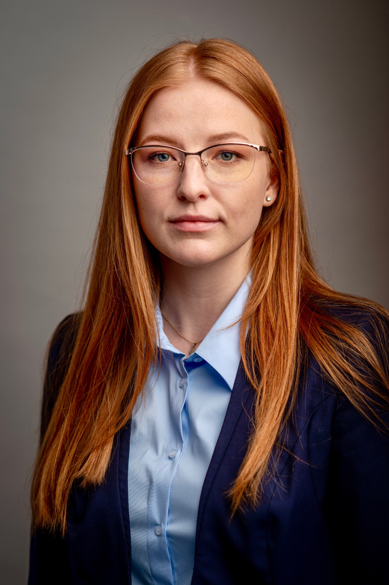 Portret kobiety z rudymi włosami w okularach i niebieskiej koszuli, ubrana w granatową marynarkę, na szarym tle. Profesjonalny wizerunek.