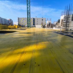 BC Budownictwo Bartłomiej Całka - Żółty szalunek stropowy na budowie w Lublinie, widok z poziomu podłogi. W tle budynki mieszkalne i żuraw wieżowy, zbrojenia. Prace w toku, słoneczny dzień.