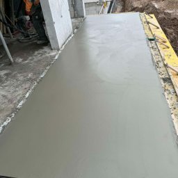 BC Budownictwo Bartłomiej Całka - Świeżo wylana, gładka betonowa płyta fundamentowa w żółtym szalunku, z widocznym pracownikiem i elementami konstrukcji budynku w tle.