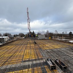BC Budownictwo Bartłomiej Całka - Zbrojenie stropu budynku w trakcie budowy, widok z góry na siatkę zbrojeniową i deskowanie, w tle pompa do betonu i pracownicy w odblaskowych kamizelkach.