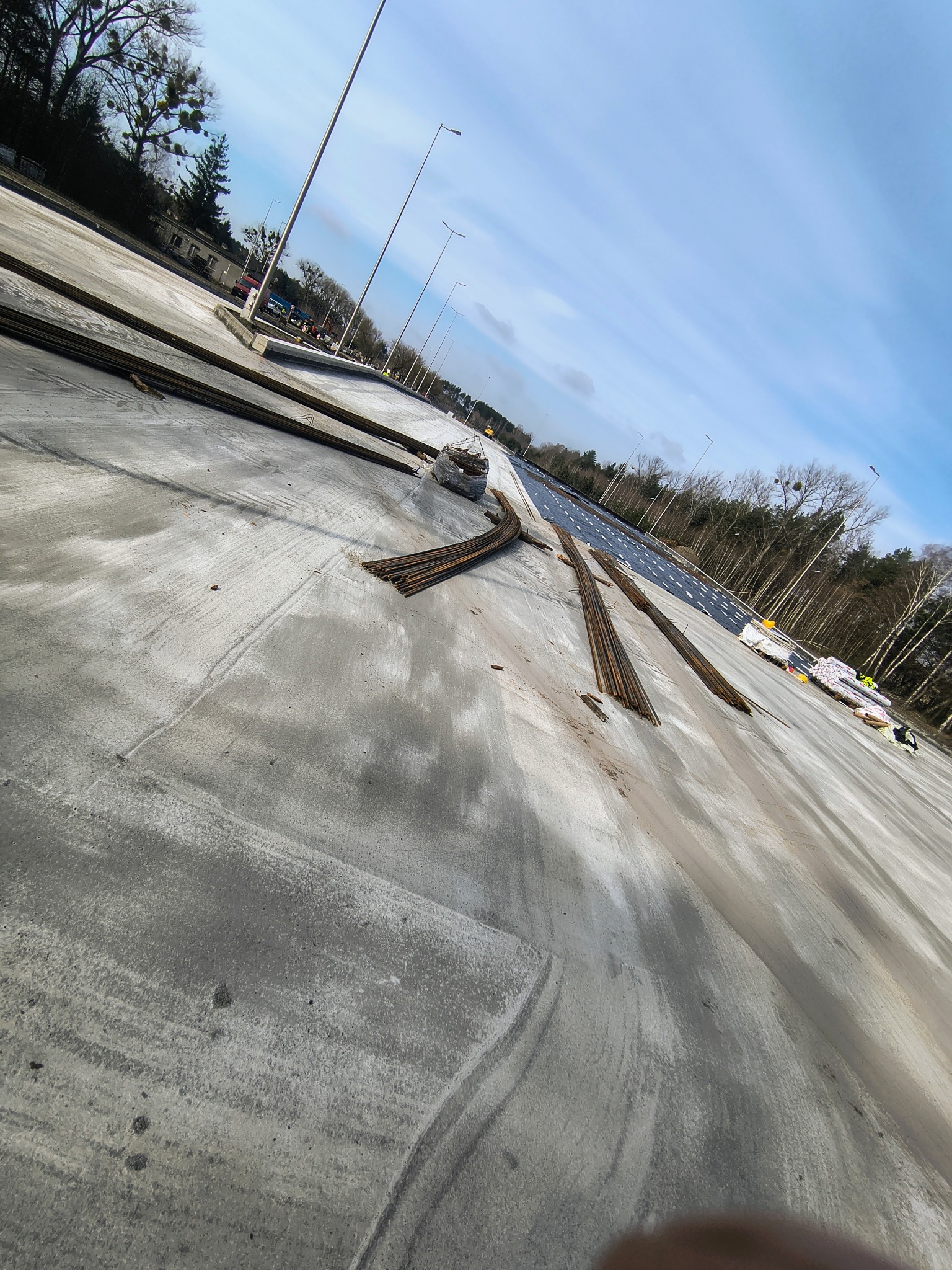 Przygotowanie zbrojenia na betonowej płycie fundamentowej pod budowę drogi, widoczne pręty zbrojeniowe i materiały izolacyjne w tle, pochmurne niebo.