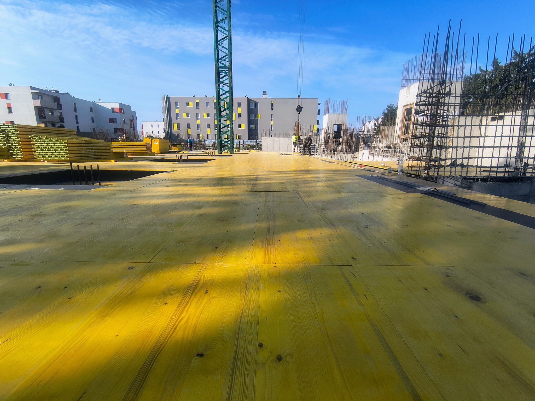 Żółty szalunek stropowy na budowie w Lublinie, widok z poziomu podłogi. W tle budynki mieszkalne i żuraw wieżowy, zbrojenia. Prace w toku, słoneczny dzień.