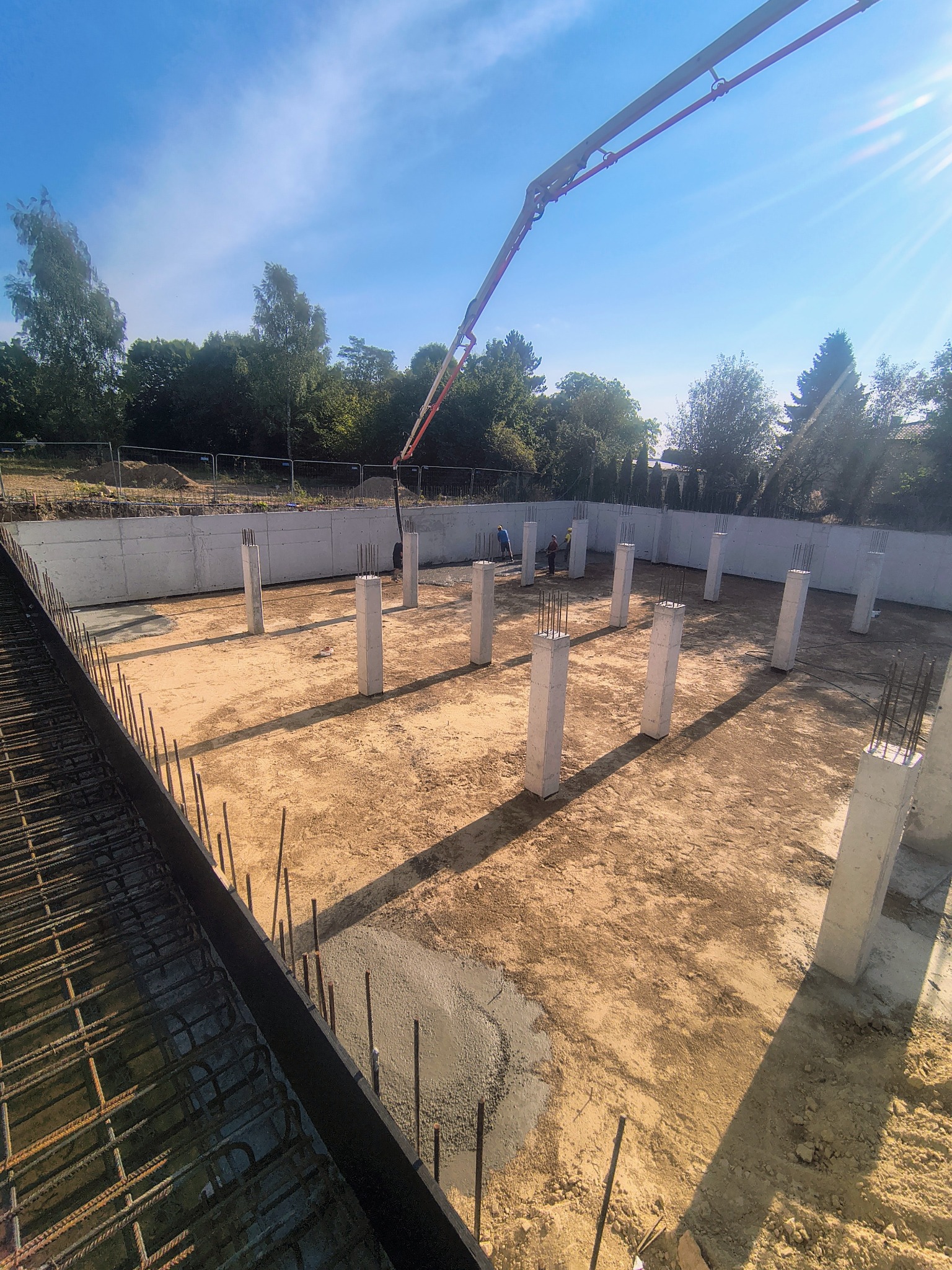 Wylewanie betonu na fundamenty pod budowę. Widoczne zbrojenia, betonowe słupy i pracownicy. Jasne niebo w tle. Prace w toku, etap budowy.