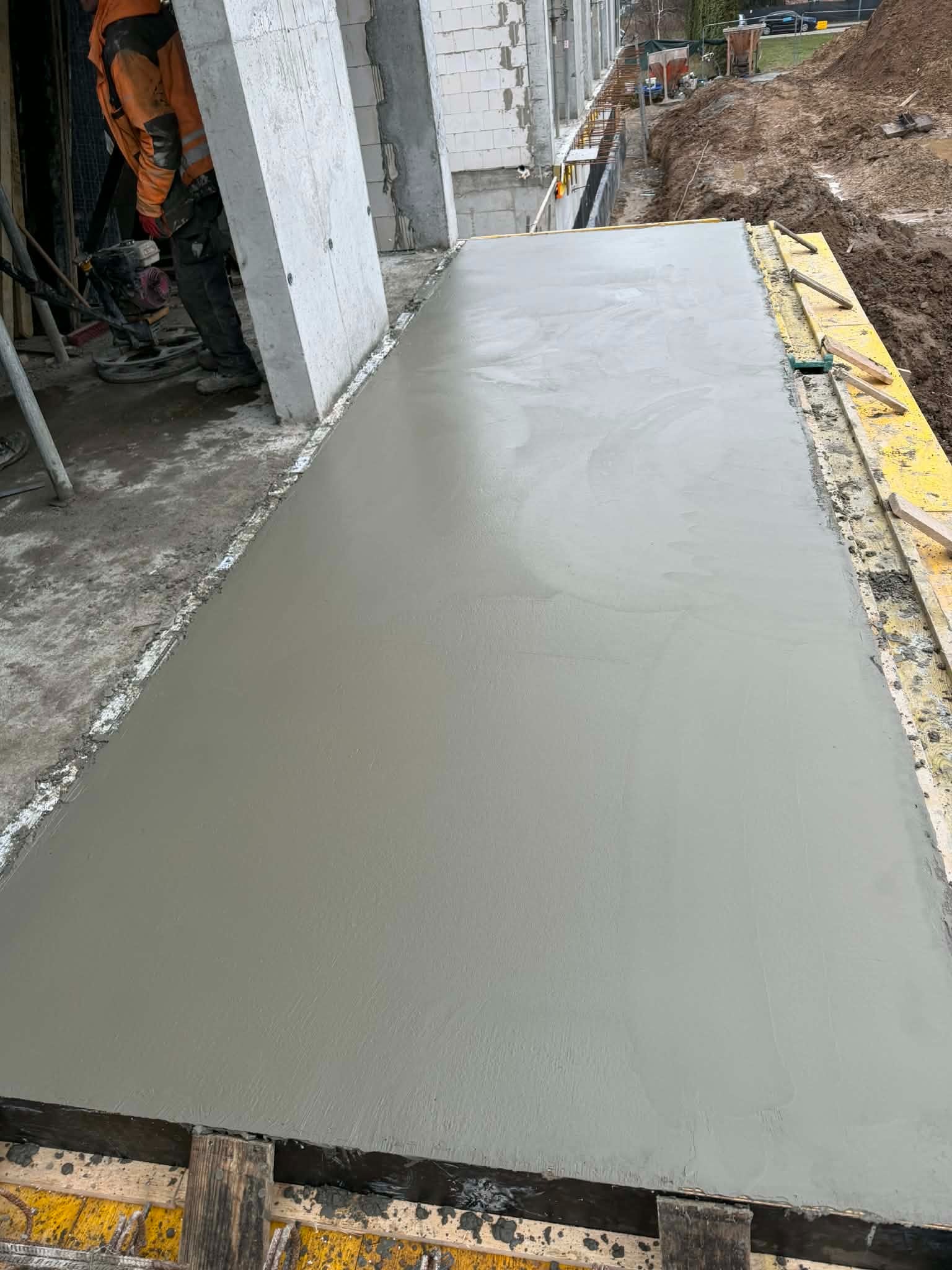 Świeżo wylana, gładka betonowa płyta fundamentowa w żółtym szalunku, z widocznym pracownikiem i elementami konstrukcji budynku w tle.