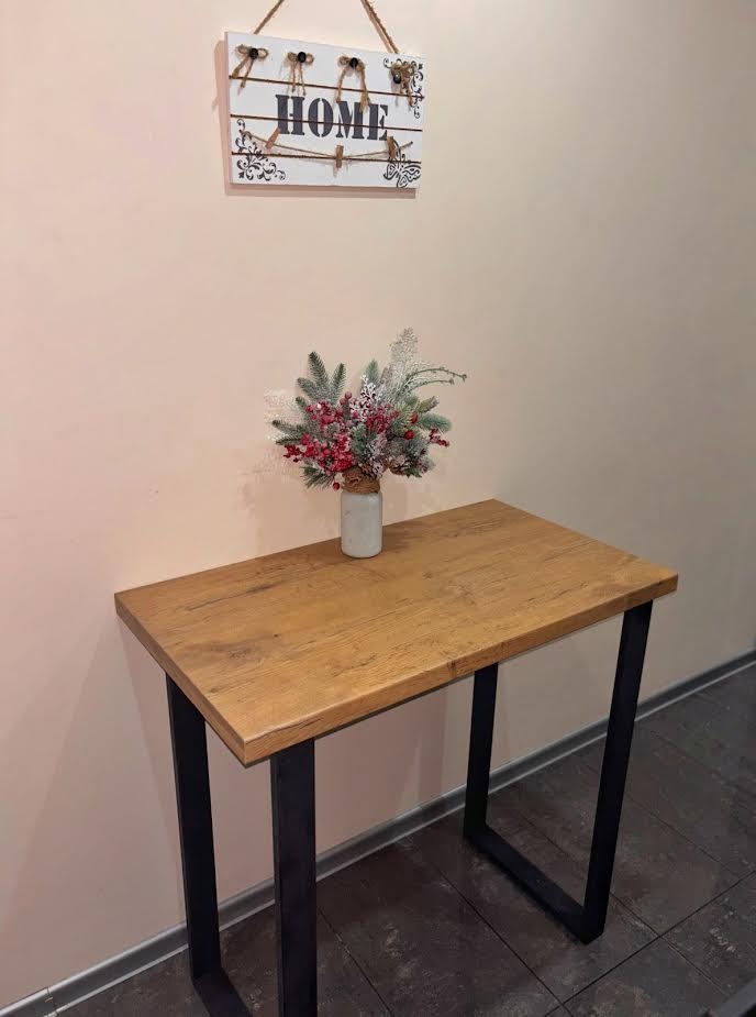 Stolik z drewnianym blatem i czarnymi, metalowymi nogami w kształcie litery U. Na blacie wazon z dekoracją świąteczną. Nad stolikiem wisi ozdobny napis 'HOME'.