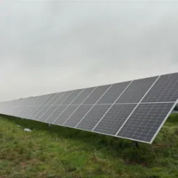 Długi rząd paneli słonecznych na zielonym polu pod pochmurnym niebem. Widoczne podpory i fragmenty instalacji. Ujęcie z niskiej perspektywy podkreśla rozległość farmy.