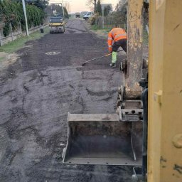 Mateusz Prusak roboty ziemno betonowe - Żwirowa droga w trakcie utwardzania walcem drogowym. Pracownik wyrównuje nawierzchnię. Widoczny fragment koparki. Praca w toku, w tle zabudowa.