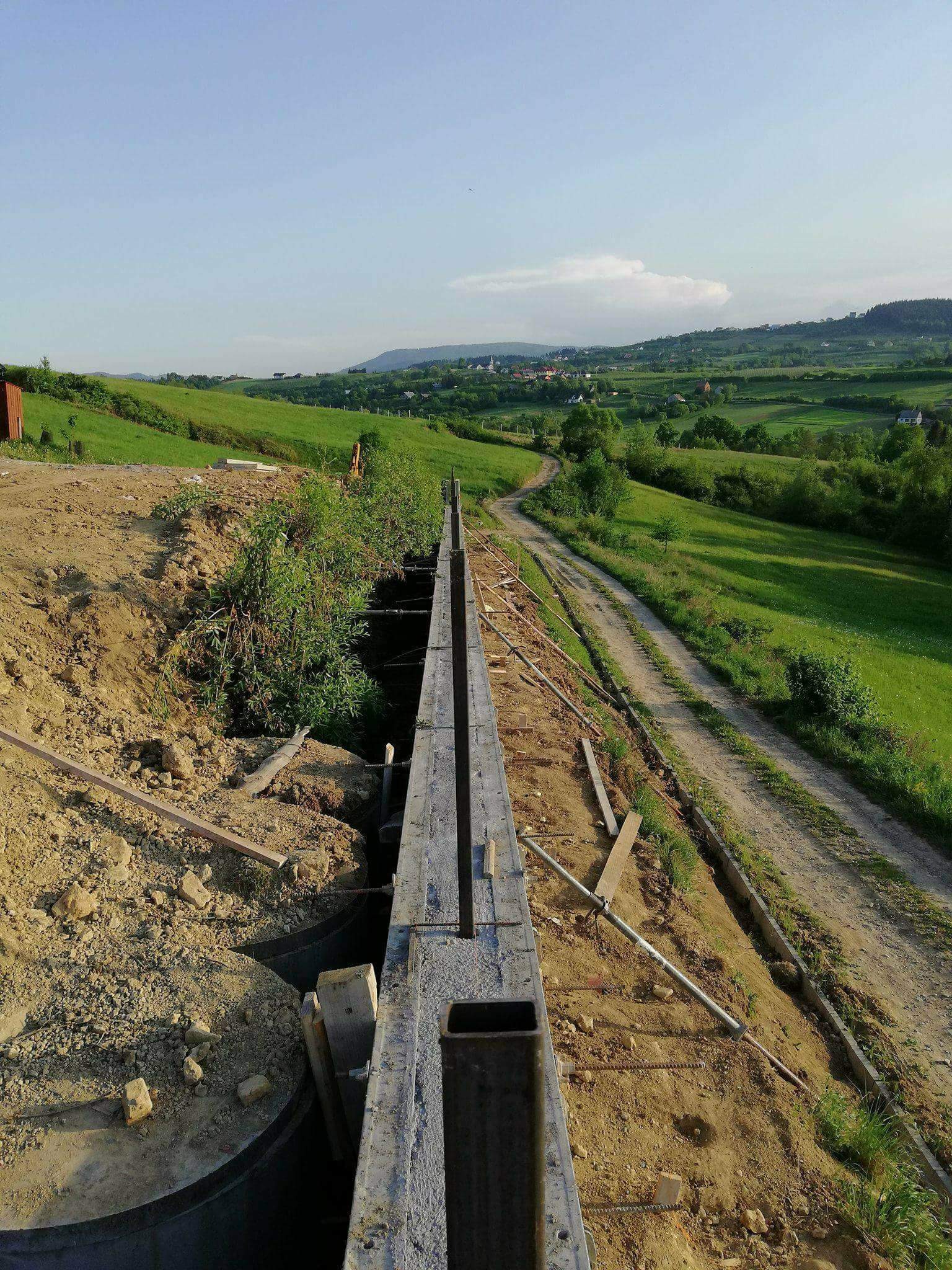 Szeroki kadr na szalunek zbrojeniowy w wykopie, z widokiem na zielone wzgórza i polną drogę w tle. Prace ziemne w toku, widoczne elementy konstrukcyjne i krajobraz.