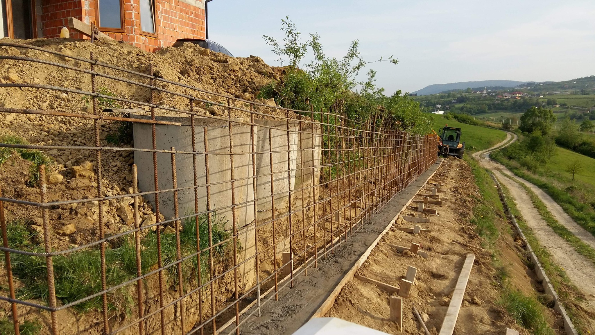 Wzmocnienie skarpy siatką zbrojeniową i betonową podmurówką. W tle traktor na drodze gruntowej i pagórkowaty krajobraz.