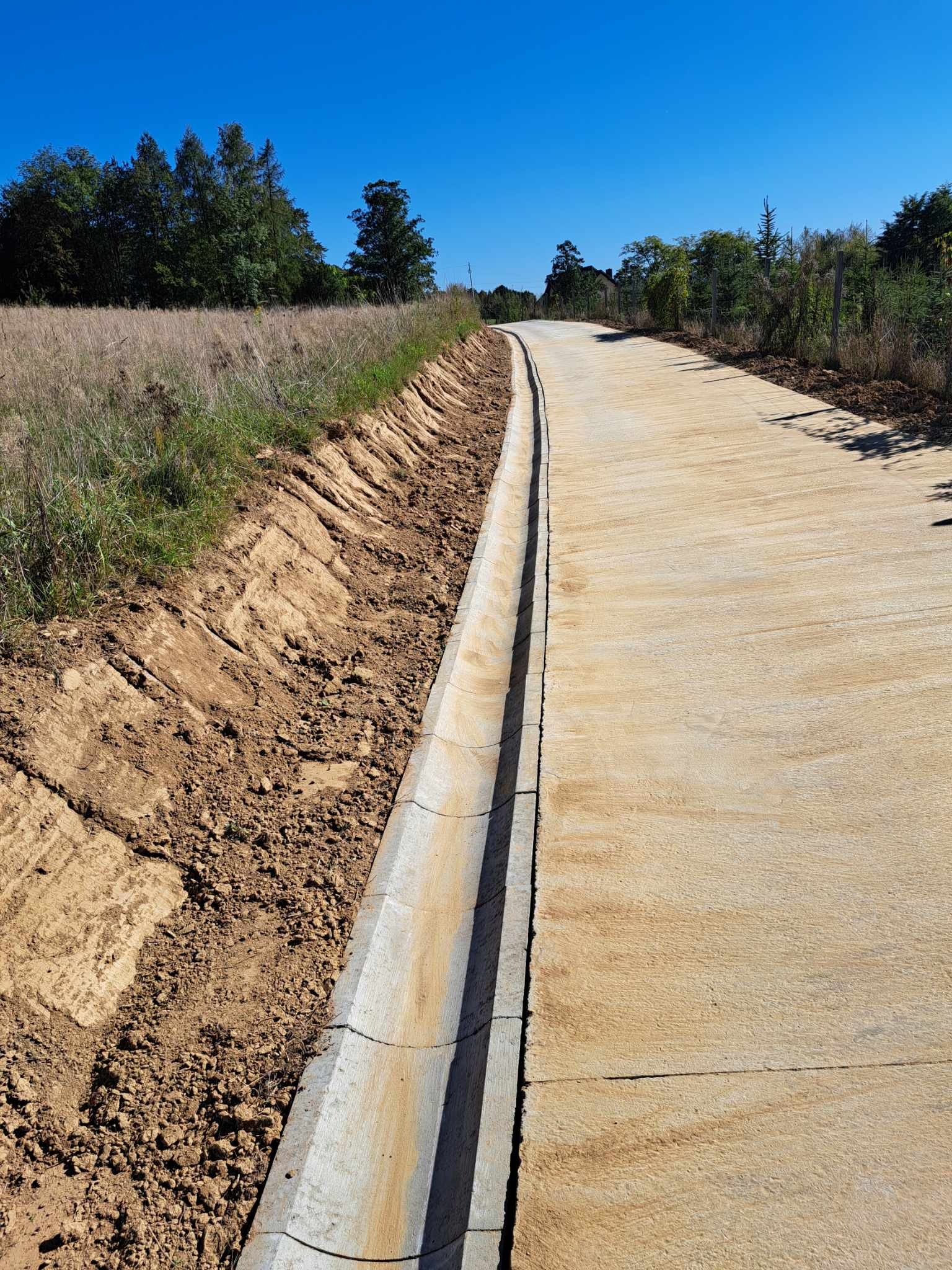 Betonowa droga z odpływem liniowym, świeżo ułożona, otoczona ziemią i trawą, pod błękitnym niebem. Widok z perspektywy ziemi, podkreślający nową infrastrukturę.