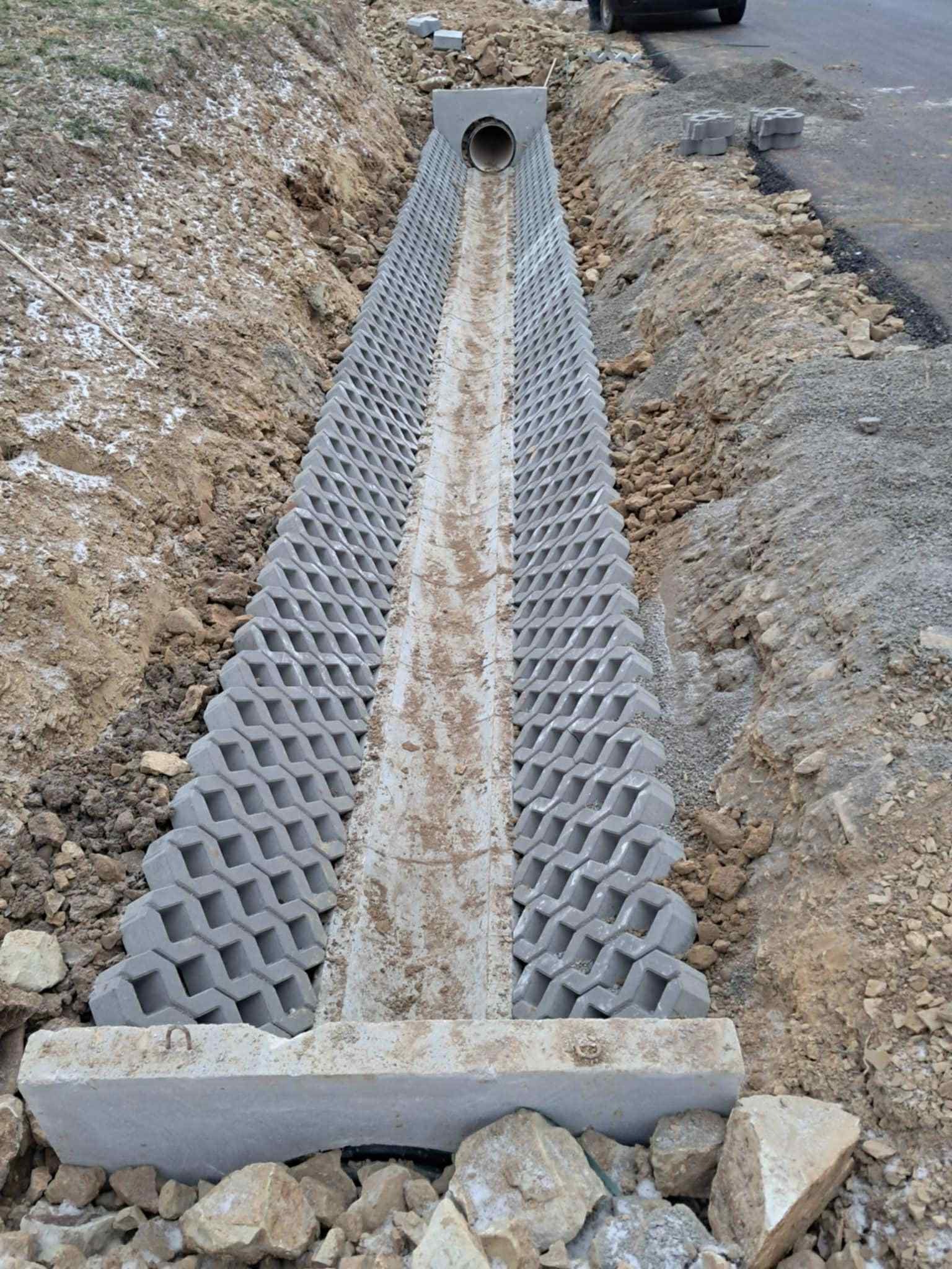 Ukończony drenaż liniowy z betonowych elementów ażurowych, odprowadzający wodę do rury w wykopie. Widoczne ułożone bloki i fragment drogi.