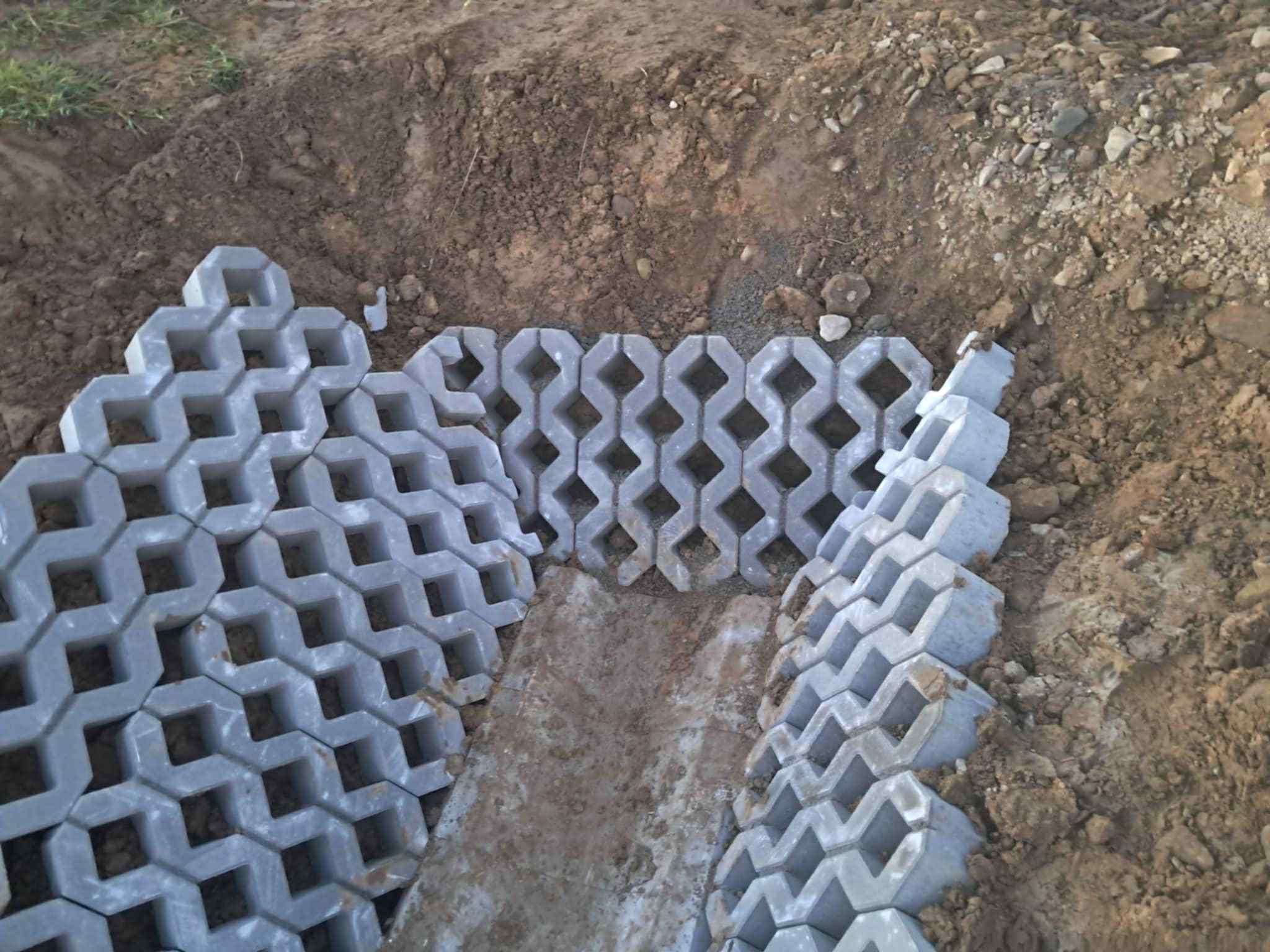 Układanie ażurowych płyt betonowych w wykopie, tworzących wzmocnienie skarpy. Widoczne fragmenty ziemi i trawy. Prace ziemne w toku.