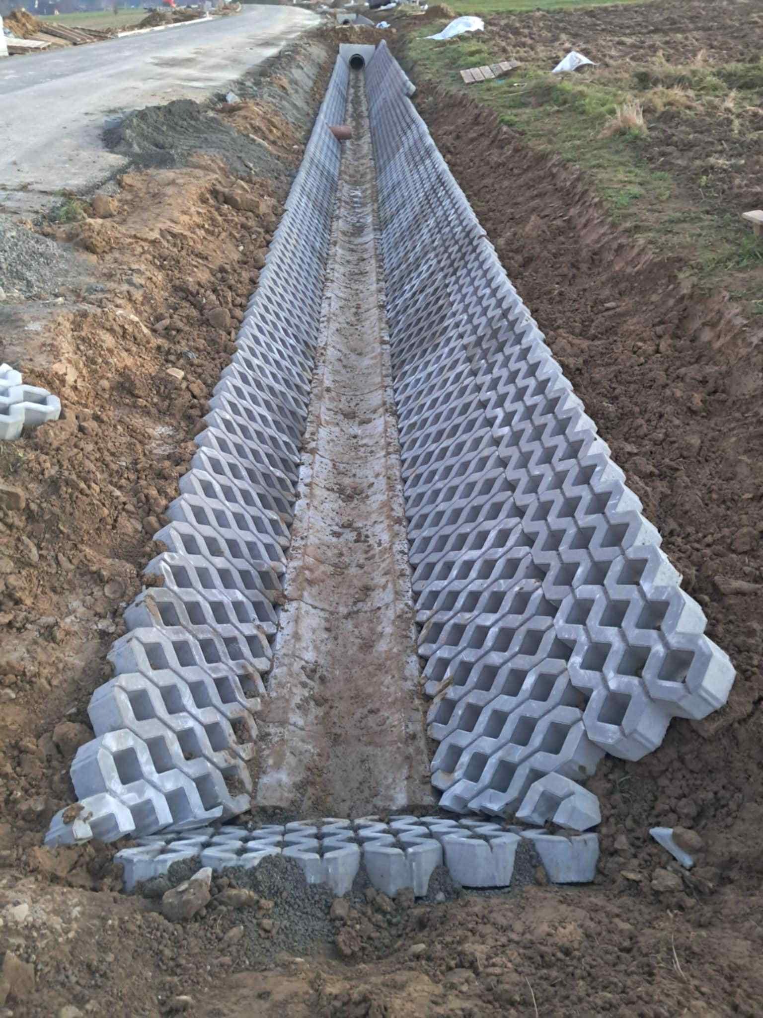 Długi, wyłożony ażurowymi betonowymi płytami rów odwadniający, biegnący wzdłuż drogi, z widoczną rurą odpływową na końcu. Ziemia po bokach, trawa w tle.