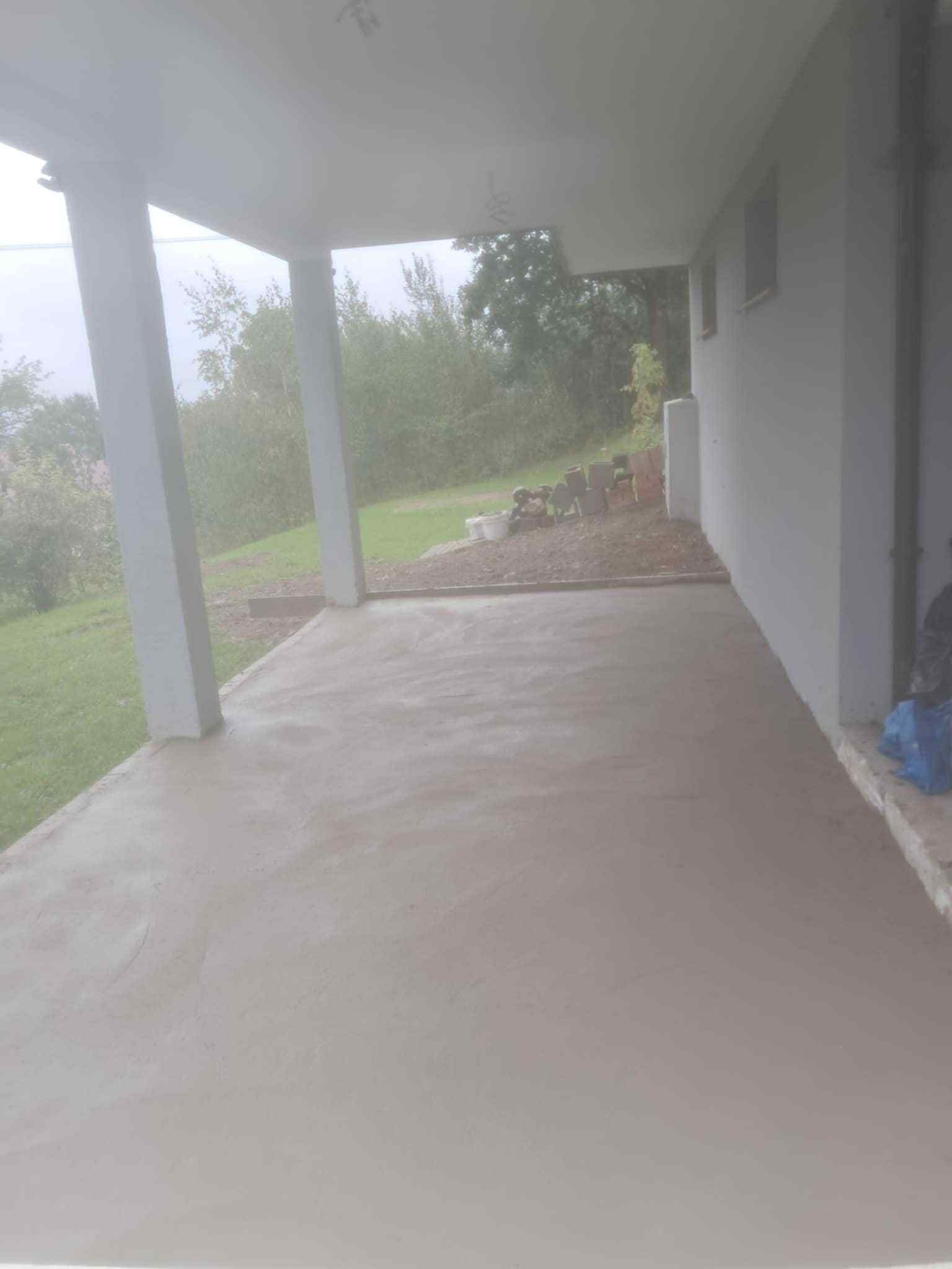 Wyrównana, świeża wylewka betonowa na zewnątrz budynku, widoczne kolumny podtrzymujące zadaszenie i fragment zieleni w tle. Prace wykończeniowe na zewnątrz.