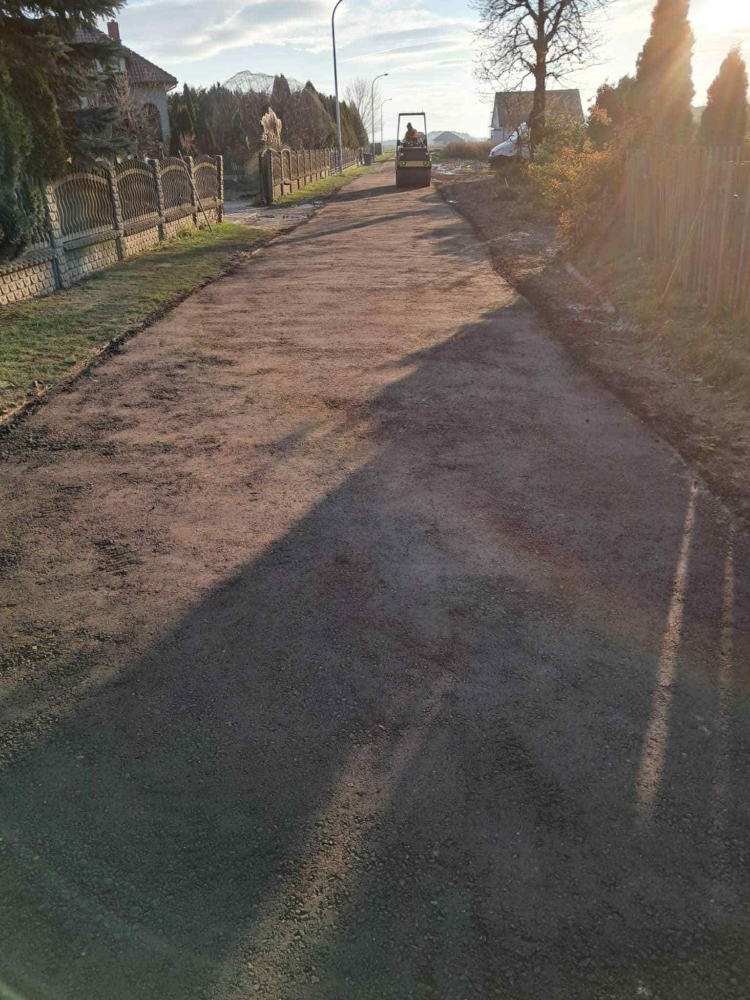 Walec drogowy ubija nawierzchnię drogi gruntowej w obszarze zabudowanym. Widok z perspektywy pieszego, w tle domy jednorodzinne i ogrodzenia.
