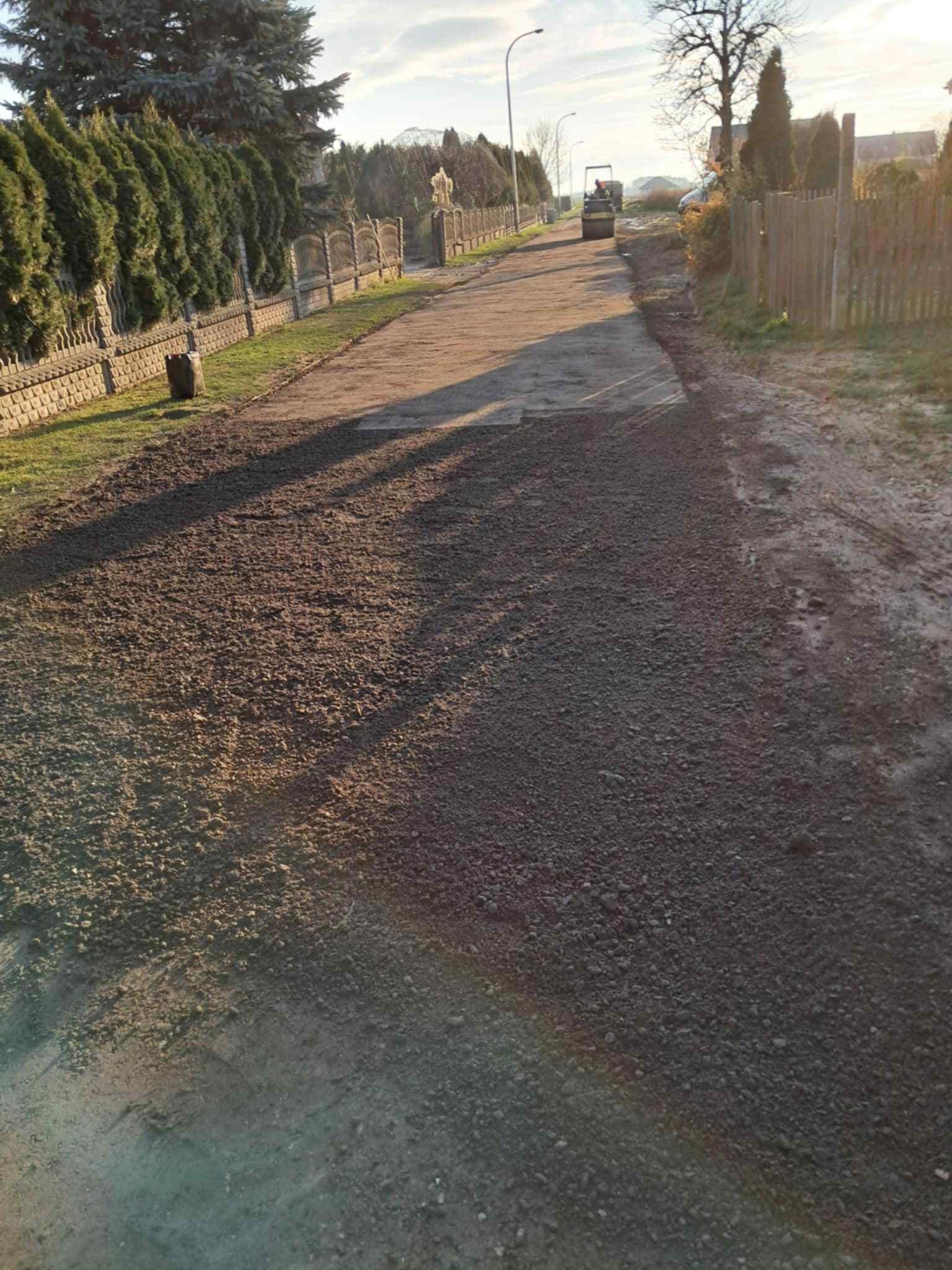 Utwardzona droga gruntowa w perspektywie, z walcem drogowym w oddali. Po lewej stronie ogrodzenie z zielenią, po prawej teren nieutwardzony. Słoneczny dzień.