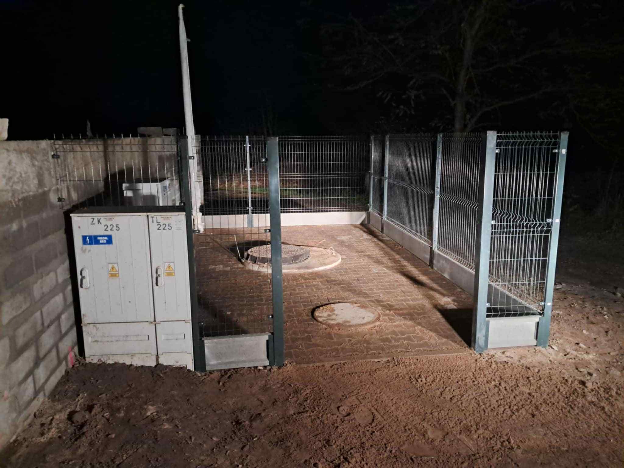 Zabezpieczone ogrodzeniem studzienki kanalizacyjne na wybrukowanym terenie, obok skrzynka elektryczna ZK 225, nocne oświetlenie.