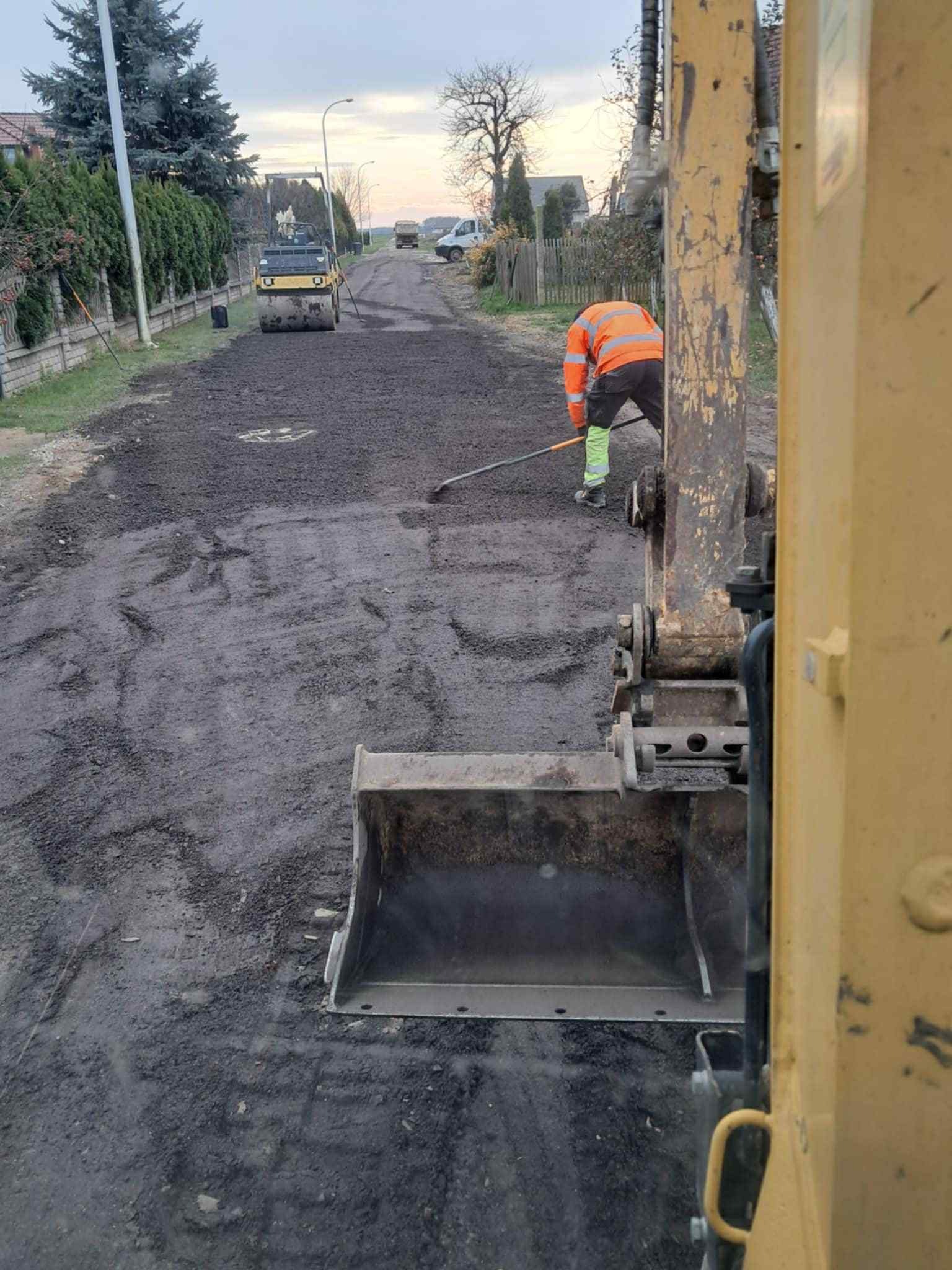 Żwirowa droga w trakcie utwardzania walcem drogowym. Pracownik wyrównuje nawierzchnię. Widoczny fragment koparki. Praca w toku, w tle zabudowa.