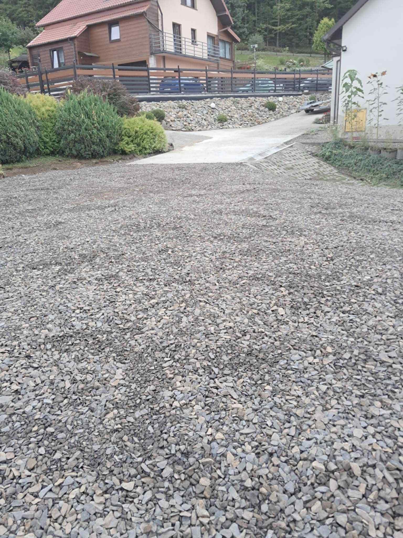 Utwardzona droga dojazdowa do domu, wysypana drobnym, szarym kamieniem. W tle widoczny dom z brązową elewacją i szarą kostką ażurową.