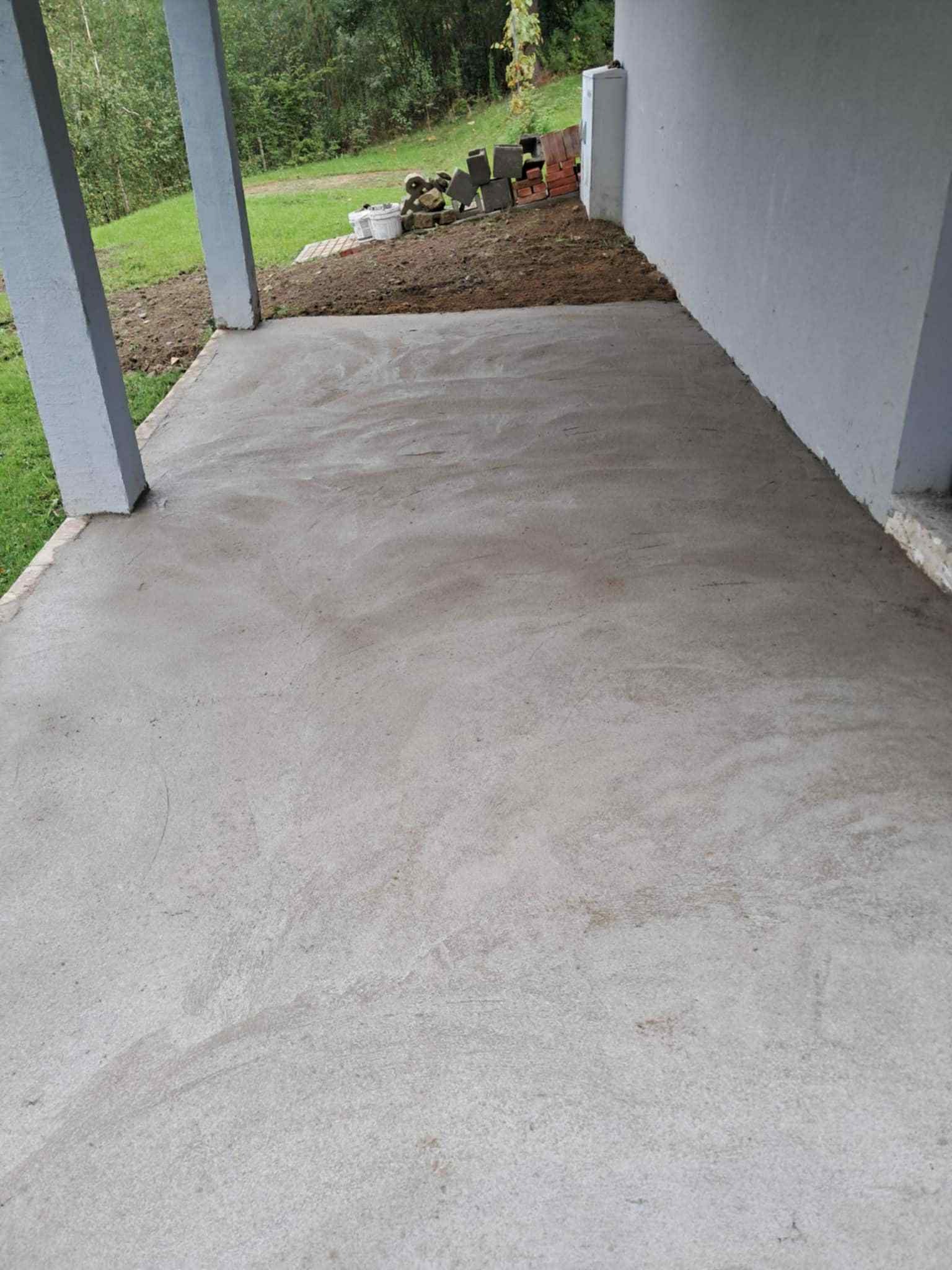 Wylana, świeża betonowa posadzka na zewnątrz budynku, widoczne podpory i fragment ściany. W tle ziemia i cegły. Szara kolorystyka betonu i elementów konstrukcyjnych.