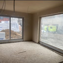 EURO-BUD - Wnętrze po nałożeniu tynków, widoczne okna z widokiem na materiały budowlane i sprzęt. Ściany gładkie, przygotowane do dalszych prac wykończeniowych.