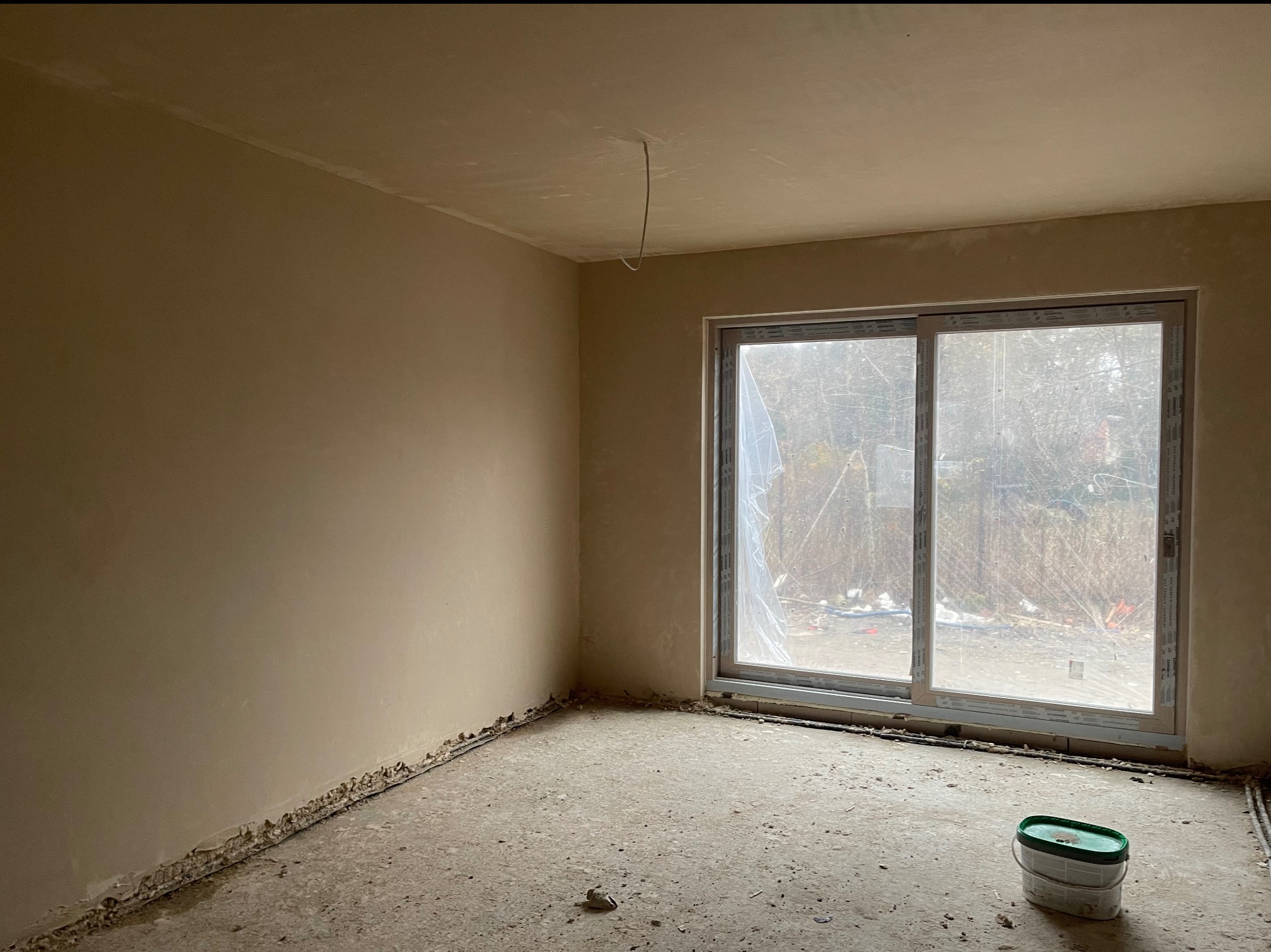 Pokój po nałożeniu tynków: gładkie, beżowe ściany i sufit, okno z widokiem na zewnątrz, wiadro na podłodze. Widoczne niedoskonałości przy podłodze.
