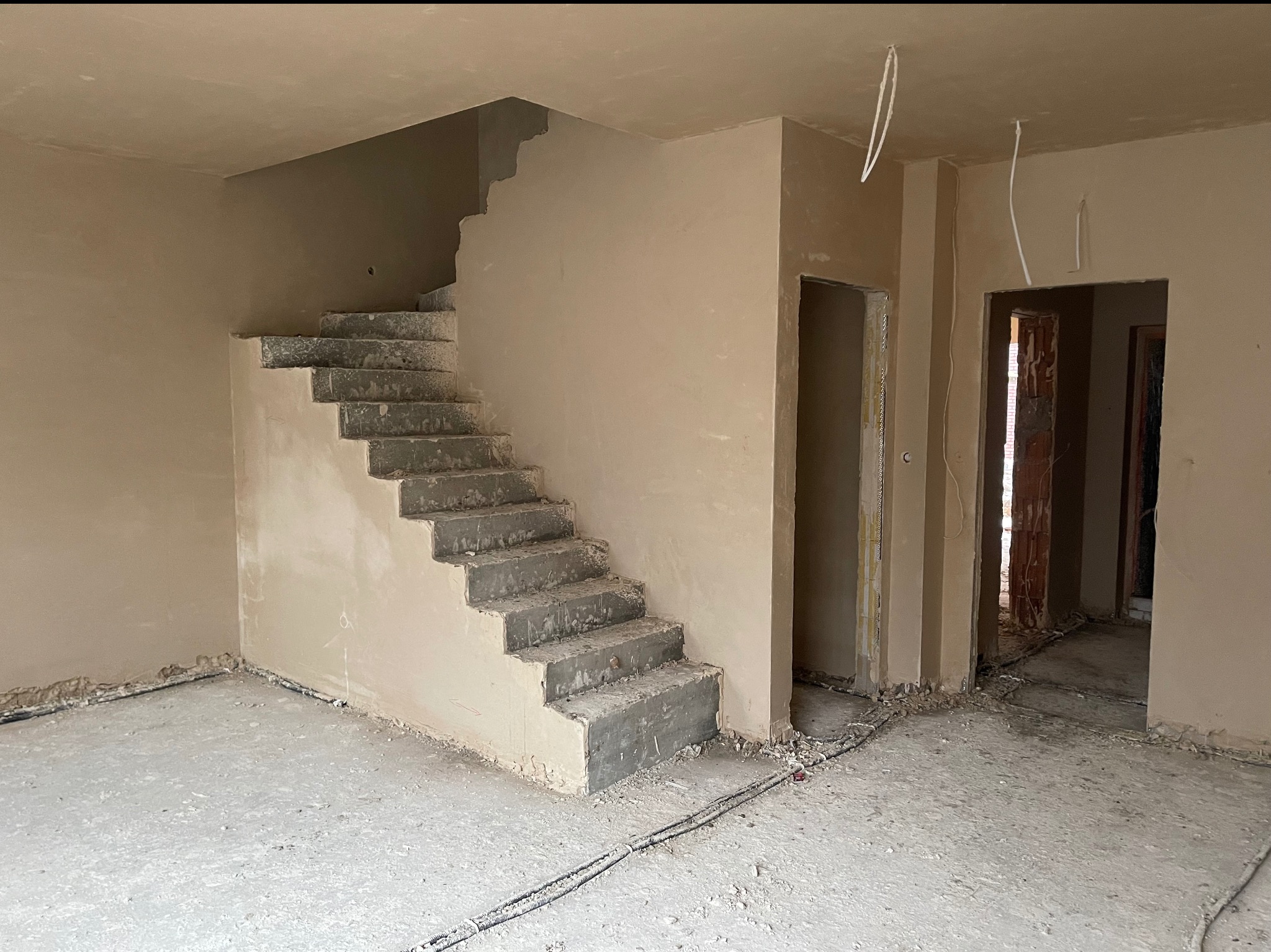 Wnętrze w trakcie tynkowania maszynowego: ściany, schody i sufit pokryte świeżym tynkiem. Widoczne kable elektryczne i surowe wykończenie otworów drzwiowych.