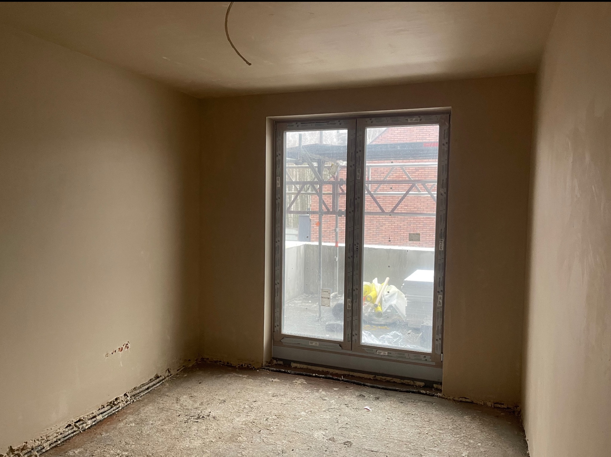 Pokój po nałożeniu tynków, widoczne gładkie ściany w beżowym odcieniu, okno z widokiem na rusztowania i budynek z czerwonej cegły. Na podłodze resztki gruzu.