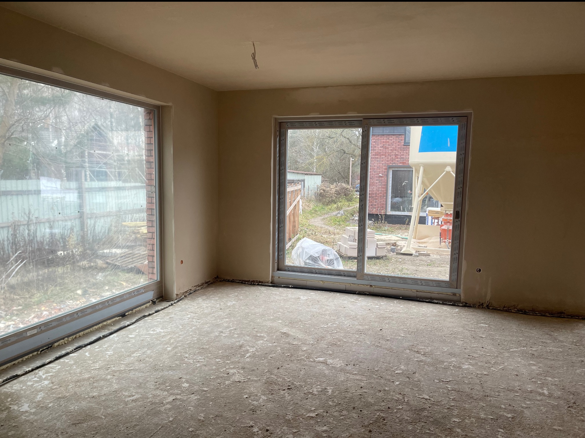 Wnętrze po nałożeniu tynku, widoczne okna z widokiem na zewnątrz, gdzie stoi maszyna do tynkowania. Ściany gładkie, podłoga surowa, przygotowana do dalszych prac.