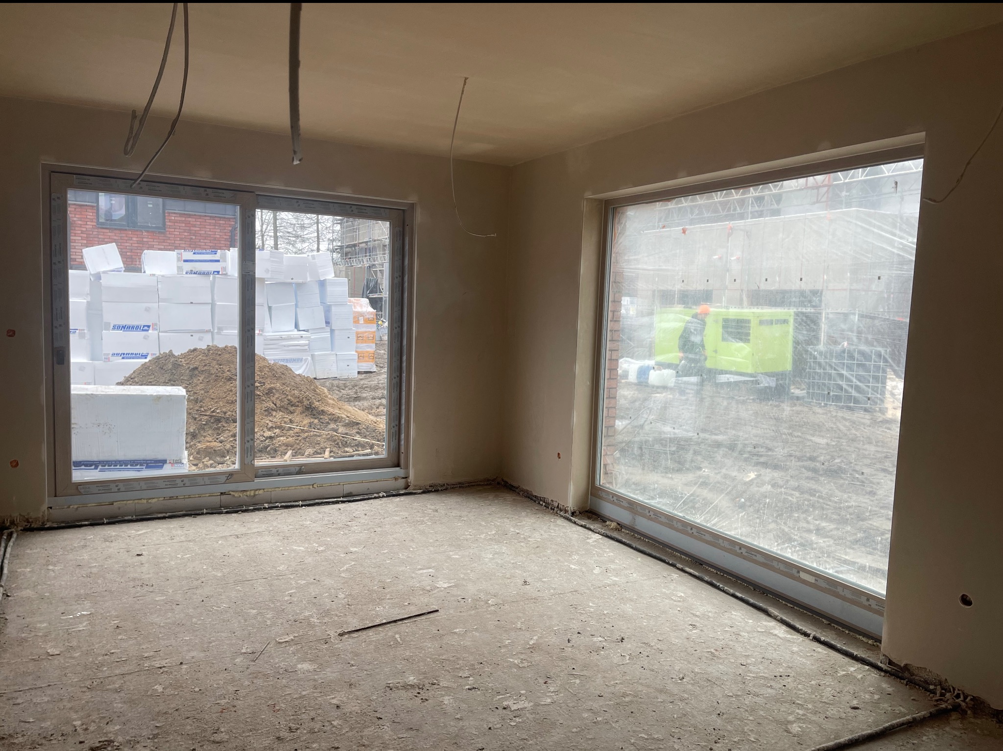 Wnętrze po nałożeniu tynków, widoczne okna z widokiem na materiały budowlane i sprzęt. Ściany gładkie, przygotowane do dalszych prac wykończeniowych.