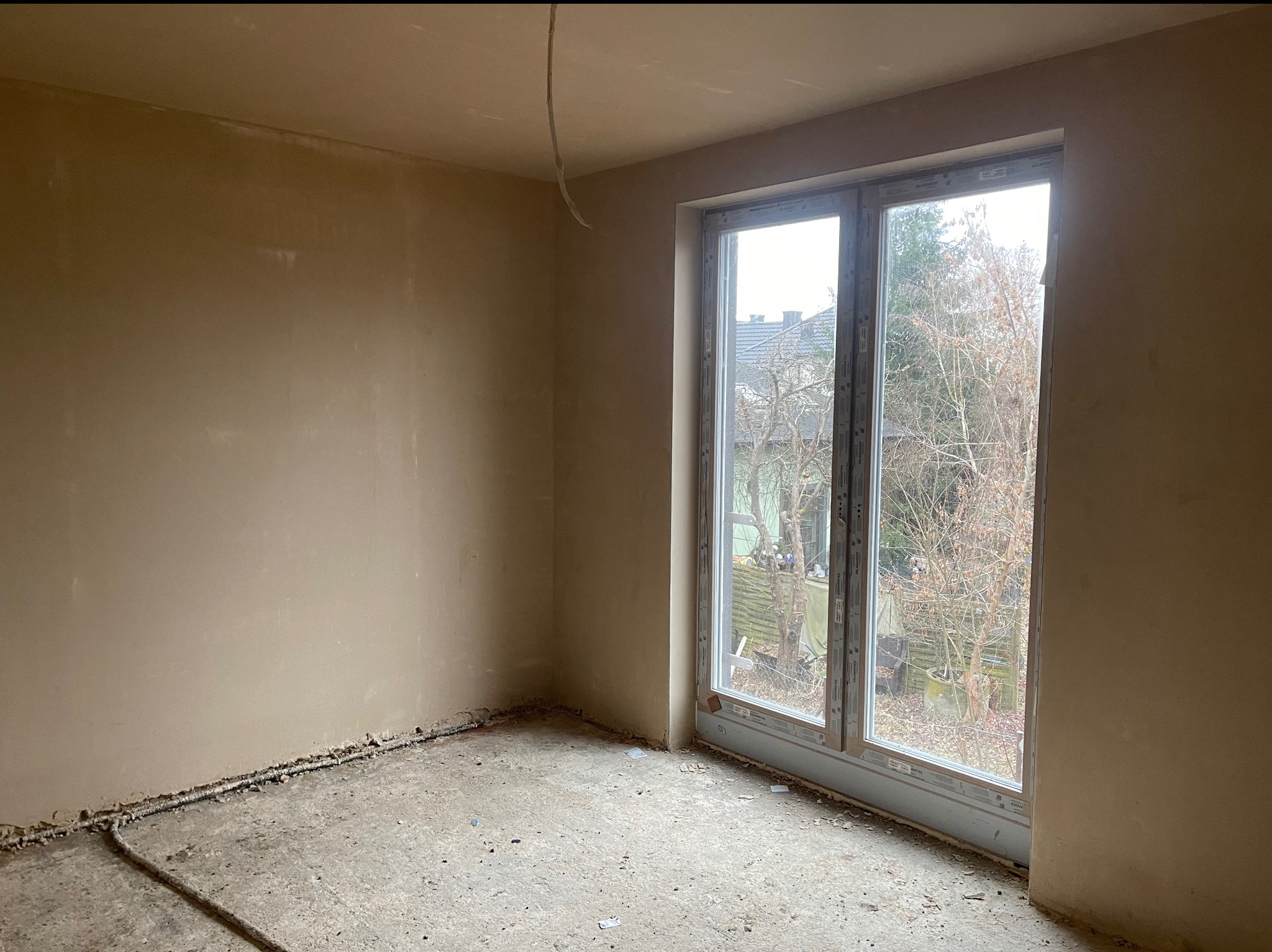 Pokój po nałożeniu tynku maszynowego, widoczne gładkie ściany w beżowym odcieniu, okno z widokiem na zewnątrz i wystające kable ze ściany.